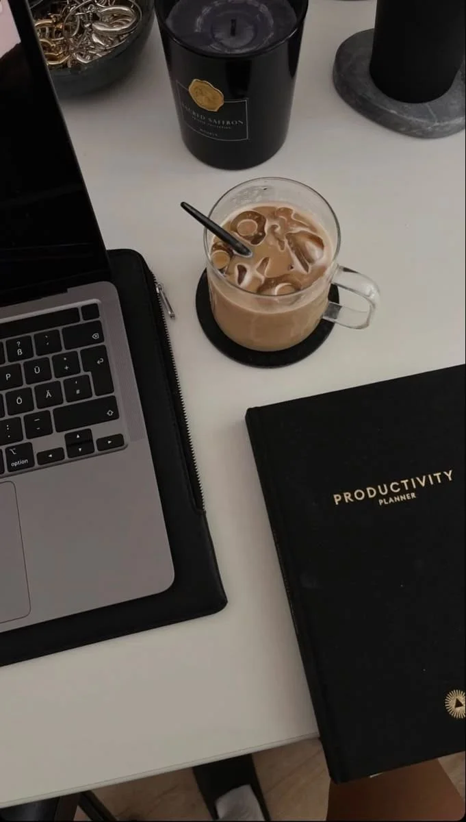 A workspace with a laptop, a black productivity planner, a glass of iced coffee, a black candle, and a bowl of jewelry on a white desk.