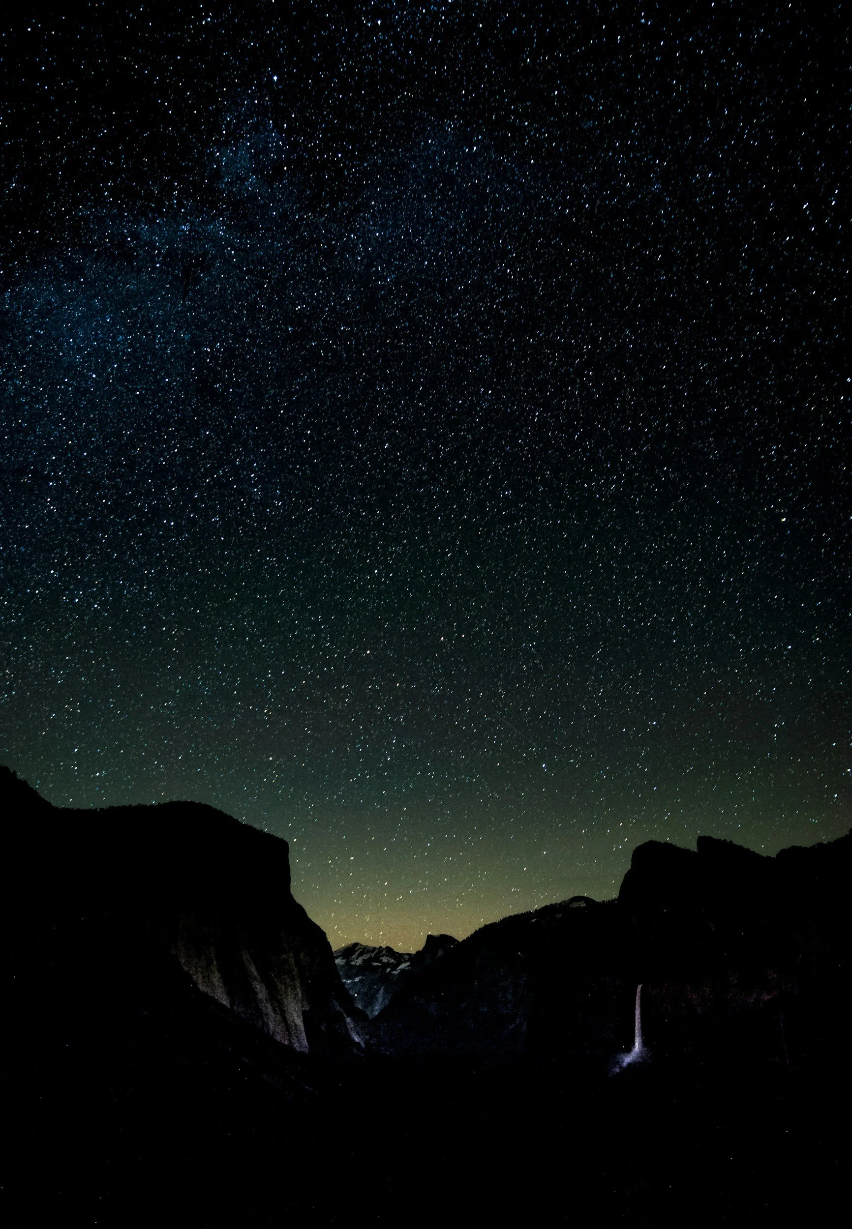 Night sky filled with numerous stars over dark mountain silhouette with a faint waterfall in a valley.