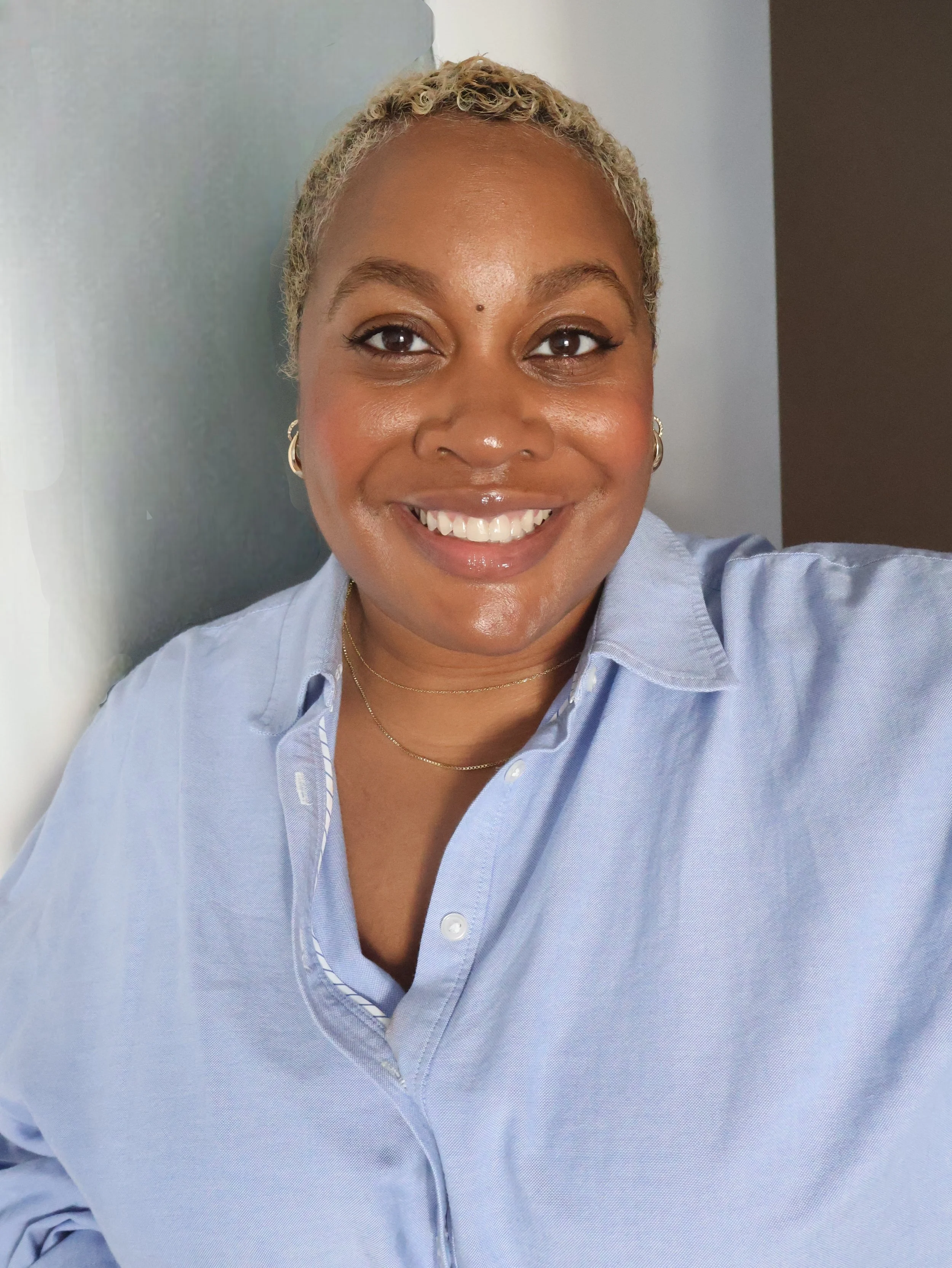 A woman with short, curly blonde hair, wearing a light blue button-up shirt and small hoop earrings, smiling at the camera.