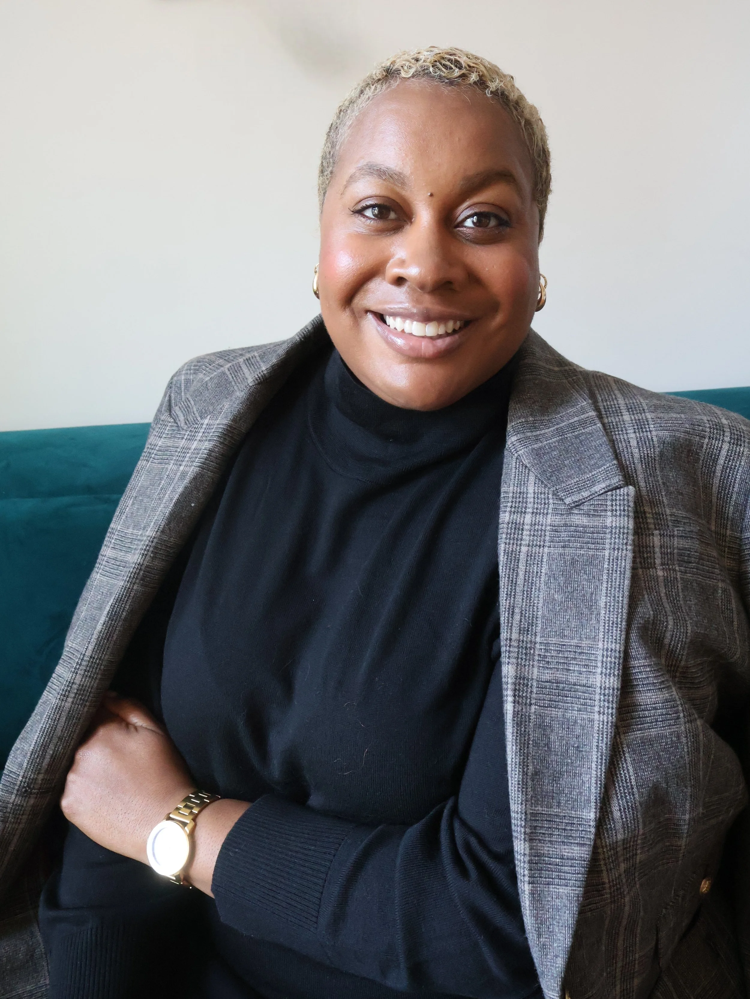 A woman smiling, wearing a black turtleneck and a plaid blazer, sitting on a teal couch.