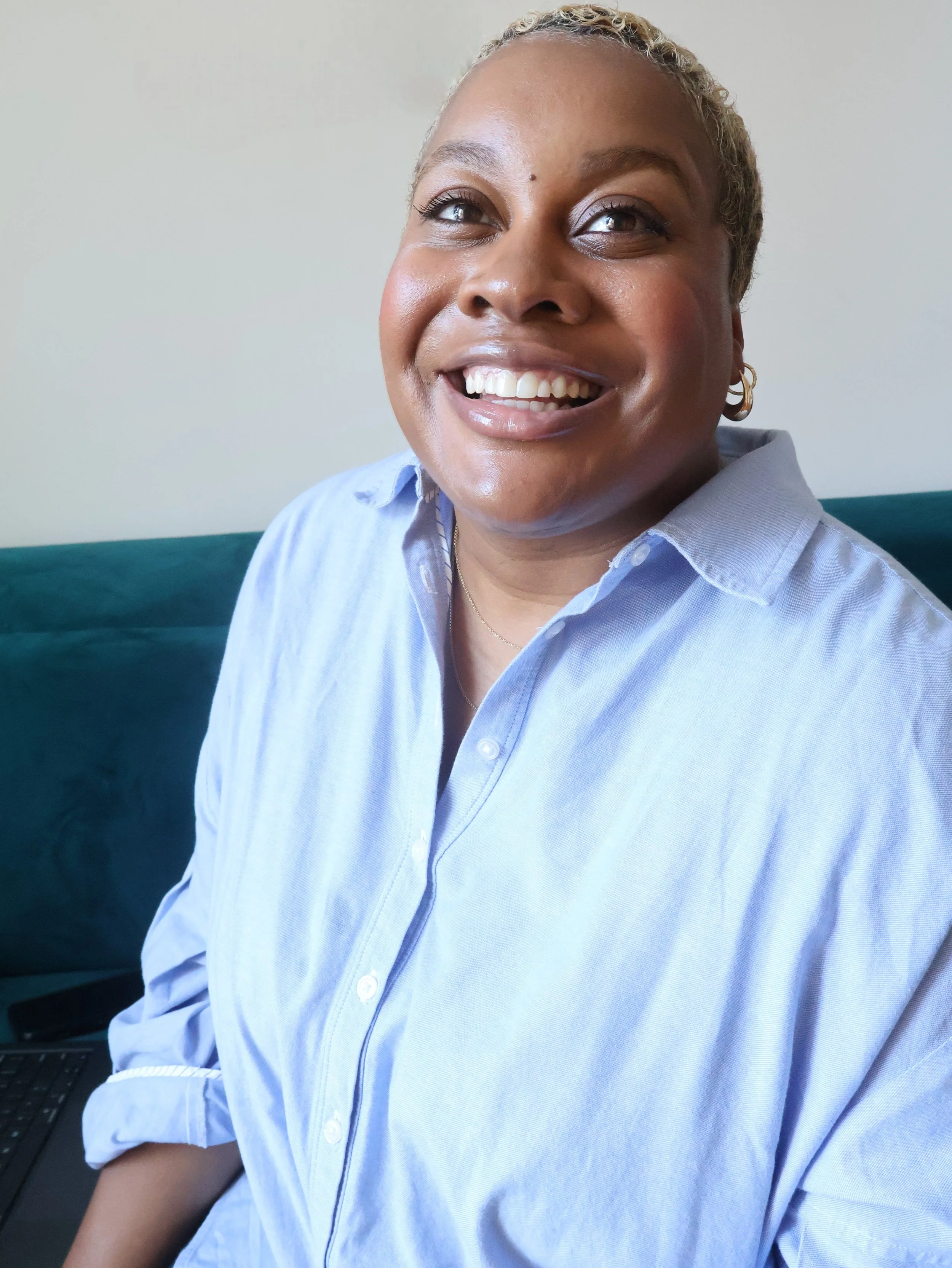 Close-up of a smiling woman with short blond hair, wearing a light blue button-down shirt, sitting indoors with a neutral background and a teal sofa.