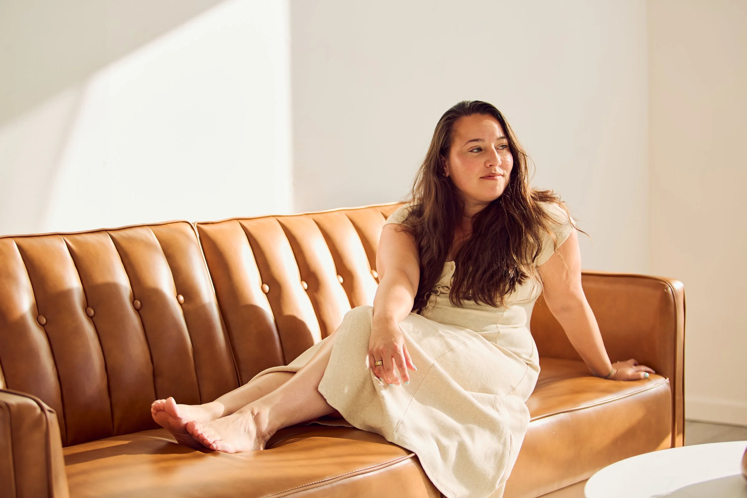 A woman with long brown hair sitting on a tan leather couch in a well-lit room.