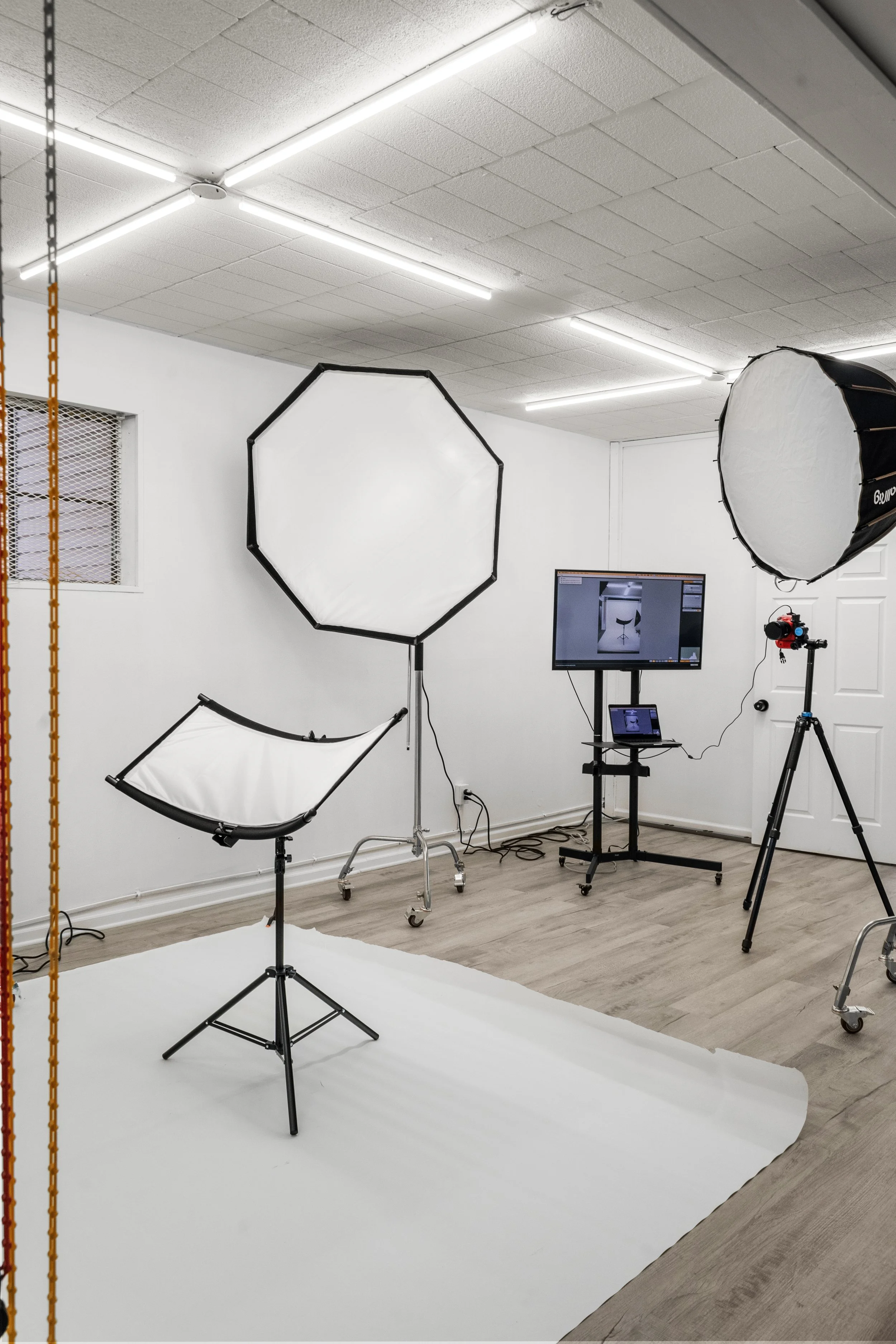 Photography studio with white backdrop, large softbox light, smaller softbox on stand, camera on tripod, TV monitor, and equipment for professional photo or video shoot.