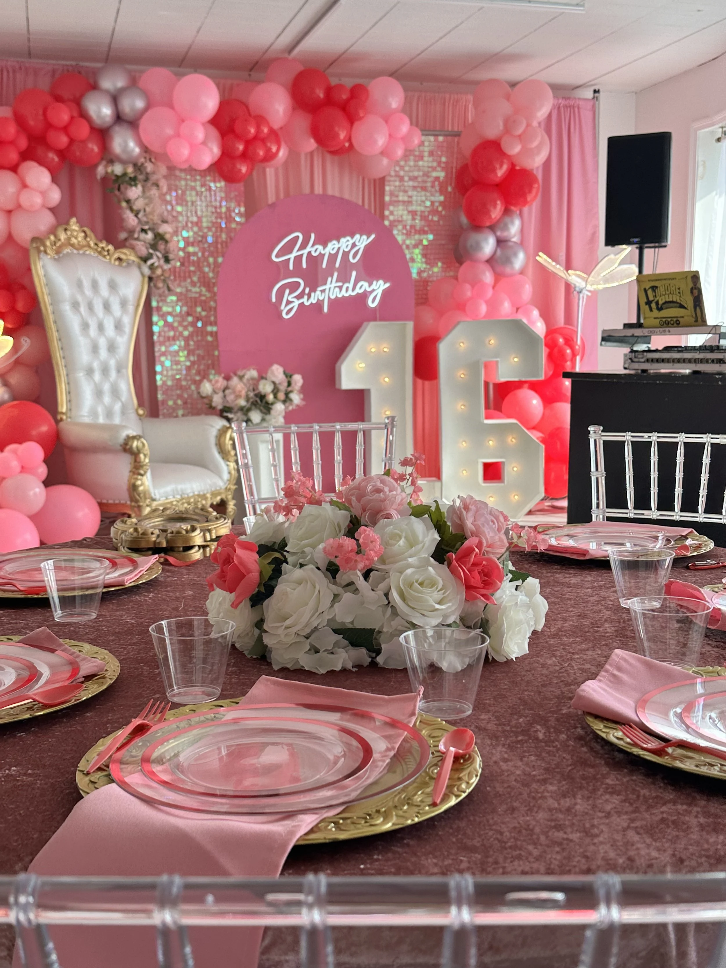 Decorated scene for a birthday celebration with a pink and red balloon arch, a large illuminated "21" sign, a white ornate chair, a round table with pink napkin and gold placemats, and a floral centerpiece. There is a pink background with "Happy Birthday" sign and a small floral arrangement.