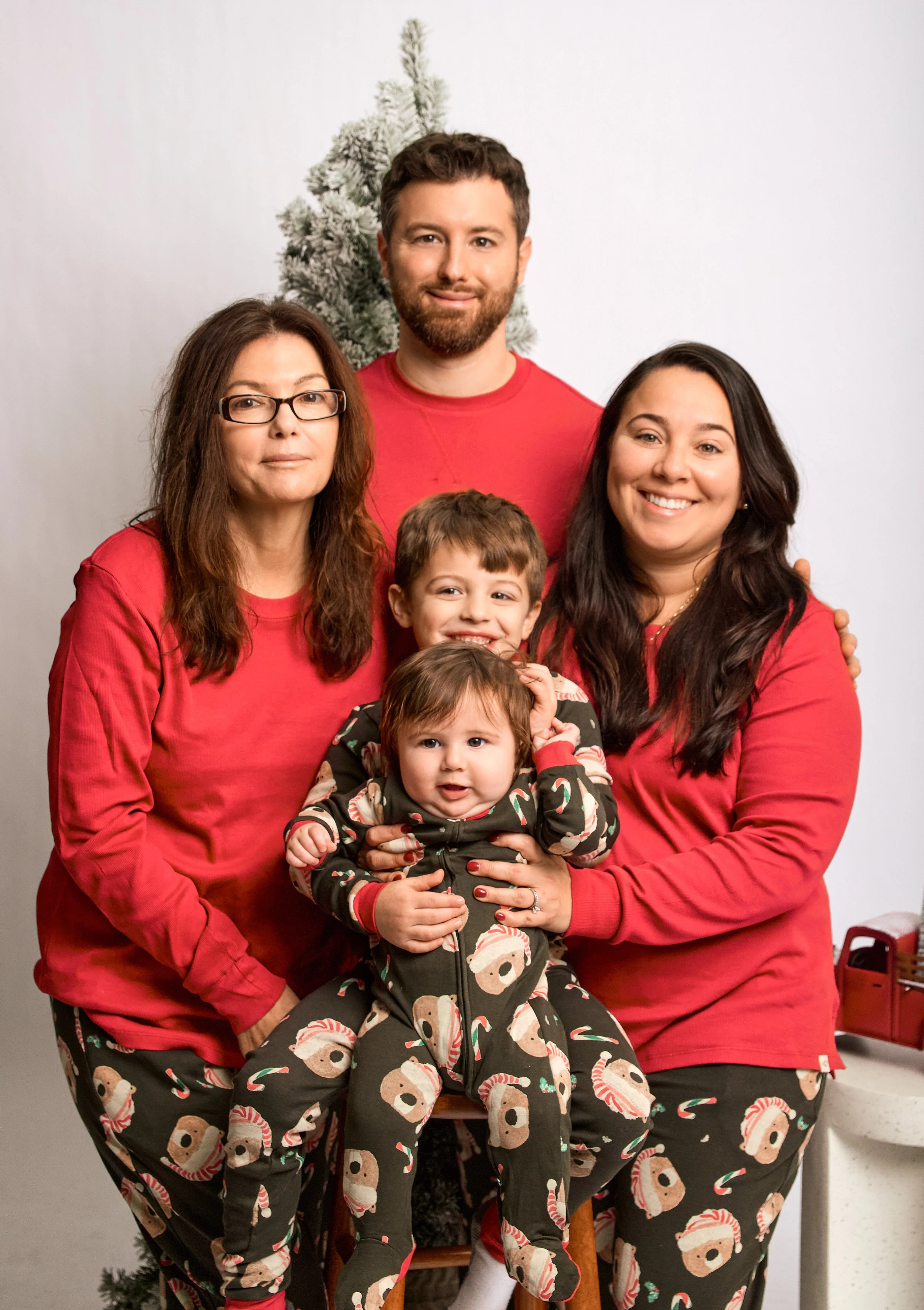 A family of six dressed in Christmas-themed pajamas and red shirts, posing together with a Christmas tree in the background. The group includes three adults and three children, all smiling.