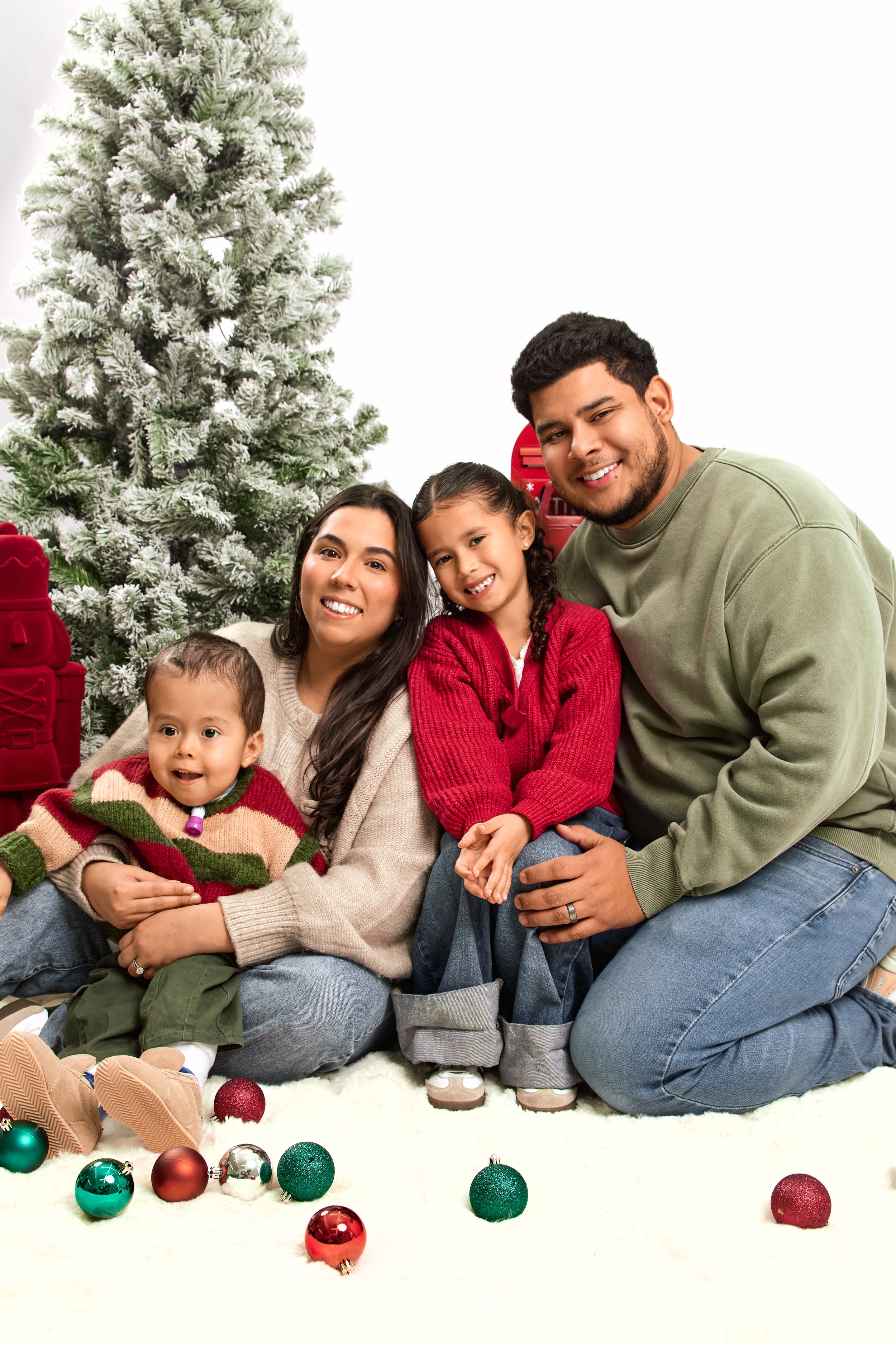 A family of four sitting on a white fluffy rug with Christmas ornaments scattered around, in front of a decorated Christmas tree.