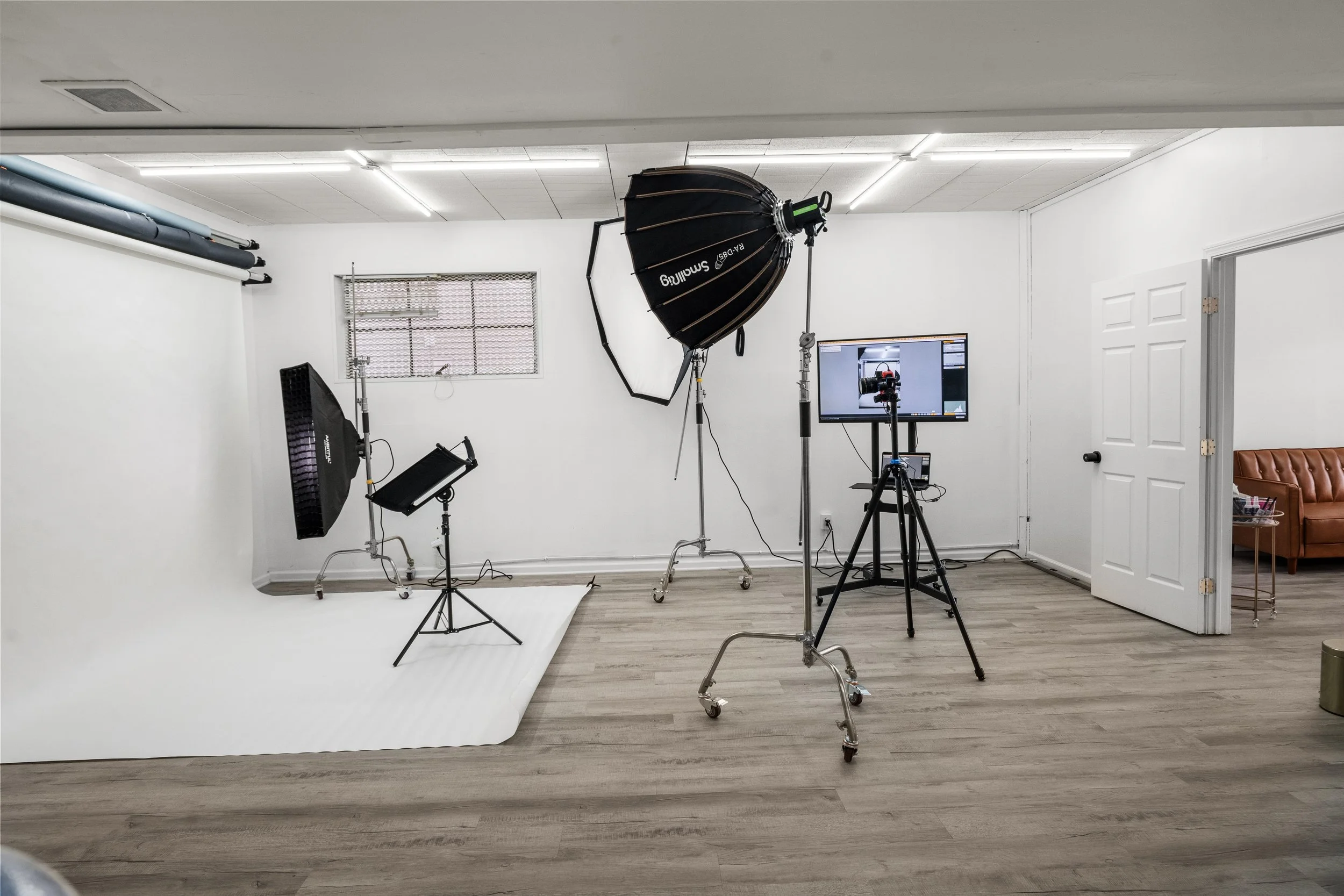 Photography studio with lighting equipment, a white backdrop, and a monitor displaying the camera view. The room has a wood floor, white walls, and a small window.