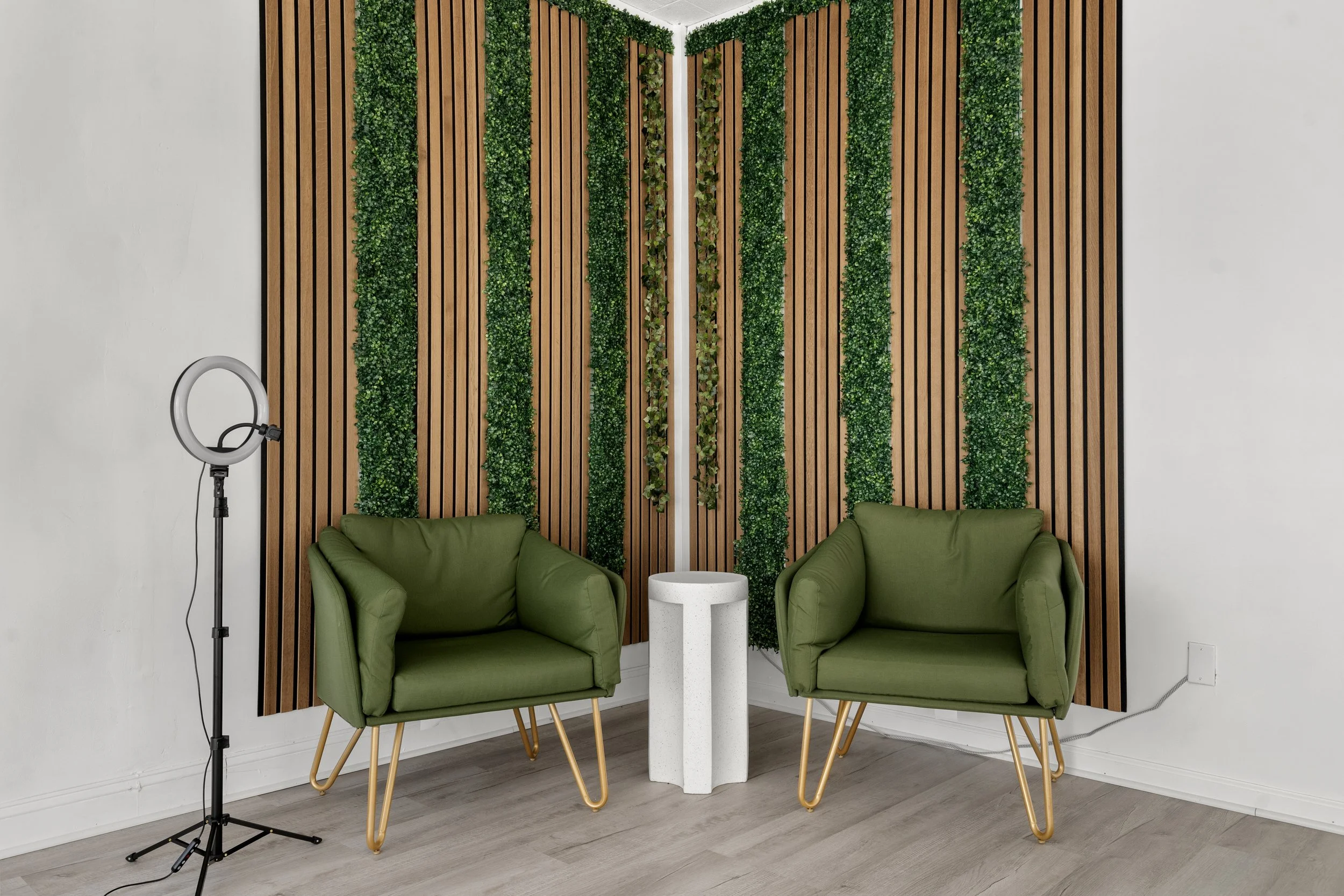 A modern interior design setup with two green armchairs with gold legs, a small rounded white table between them, a vertical wooden slat wall with green foliage accents, and a ring light on a stand to the left, all on light wood flooring.