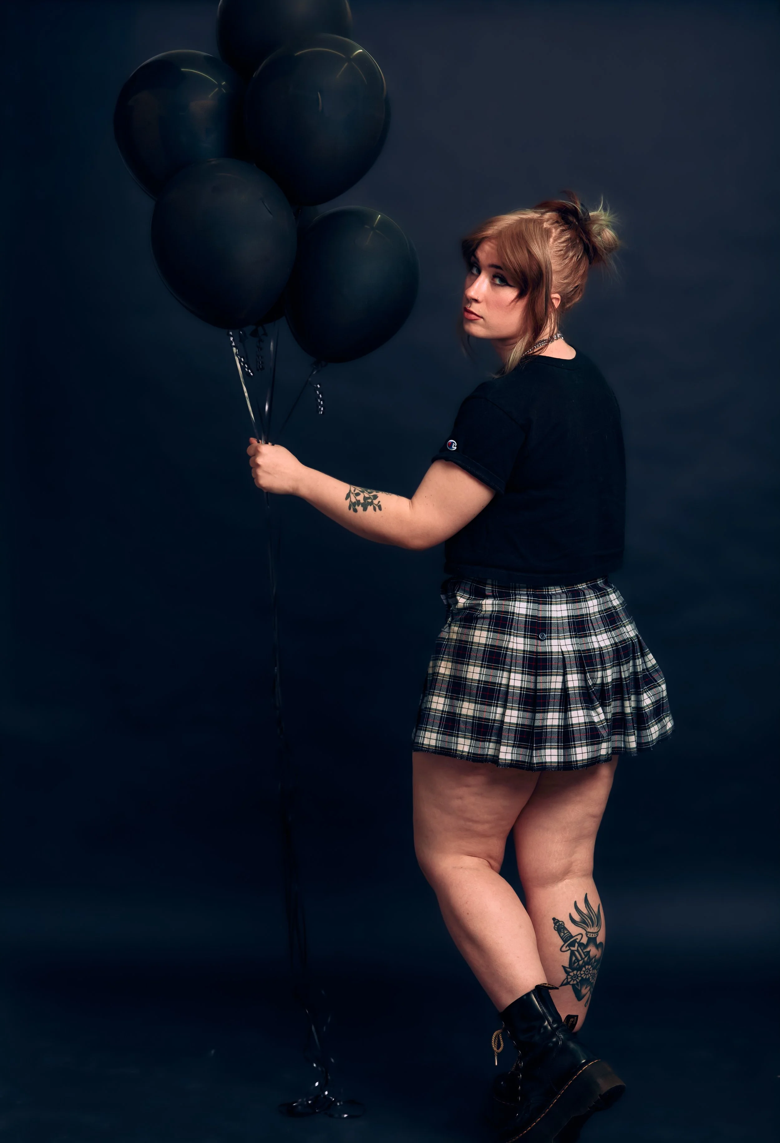 A woman with red hair, wearing a black t-shirt, plaid skirt, and black boots, holding five black balloons against a dark background.