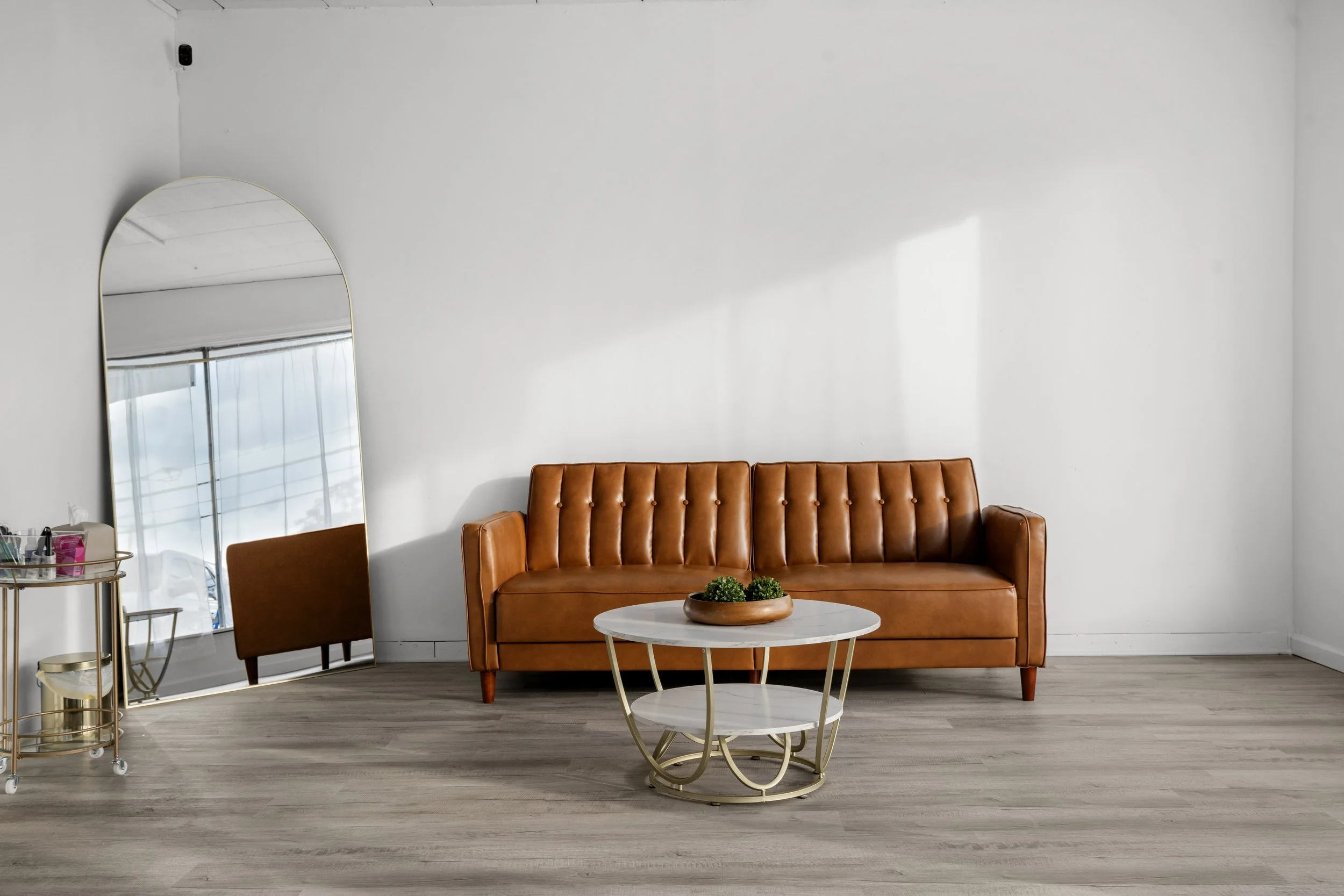 Studio room with a brown leather sofa, a round marble-top coffee table with a gold frame, a large mirror on the left, and a cart with beauty products, against white walls and gray wood flooring.
