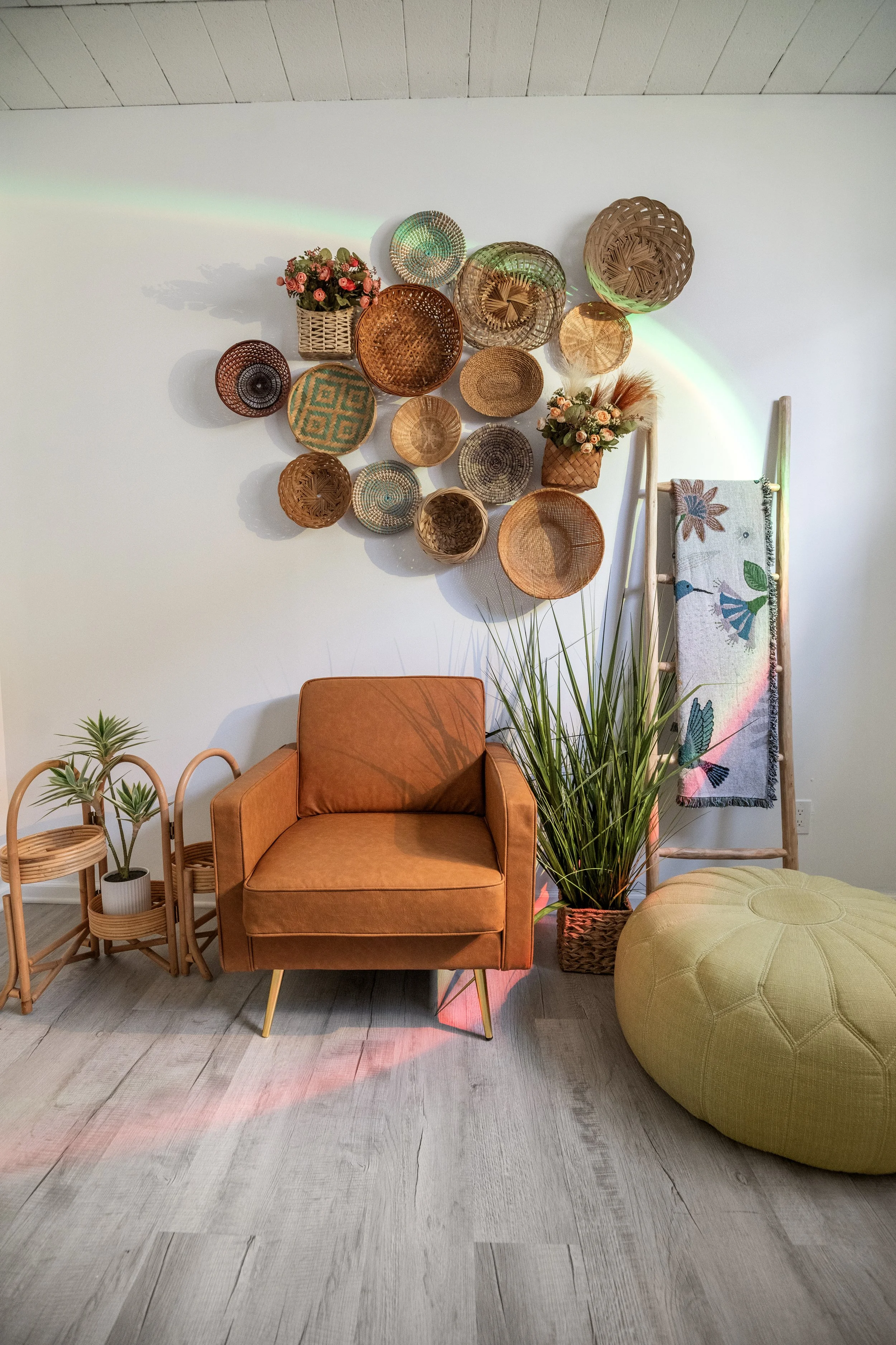 Decorative studio room corner with a brown armchair, green plants, woven baskets on the wall, colorful woven blanket, and a large yellow ottoman.