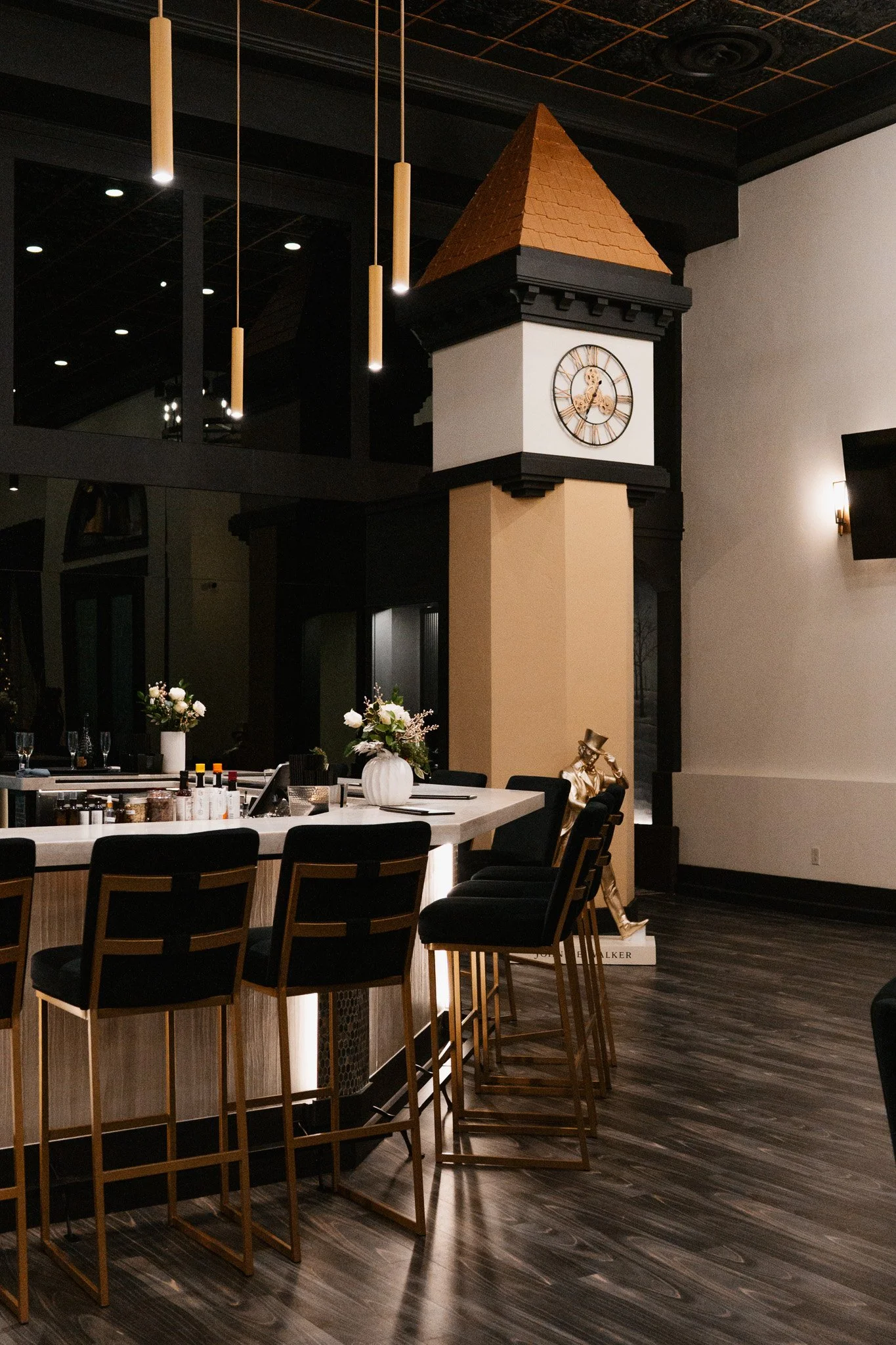 Interior of a modern restaurant or bar with a tall clock tower, black chairs with gold frames, a white marble counter with flowers, and a gold statue of a man in a hat standing on a book.