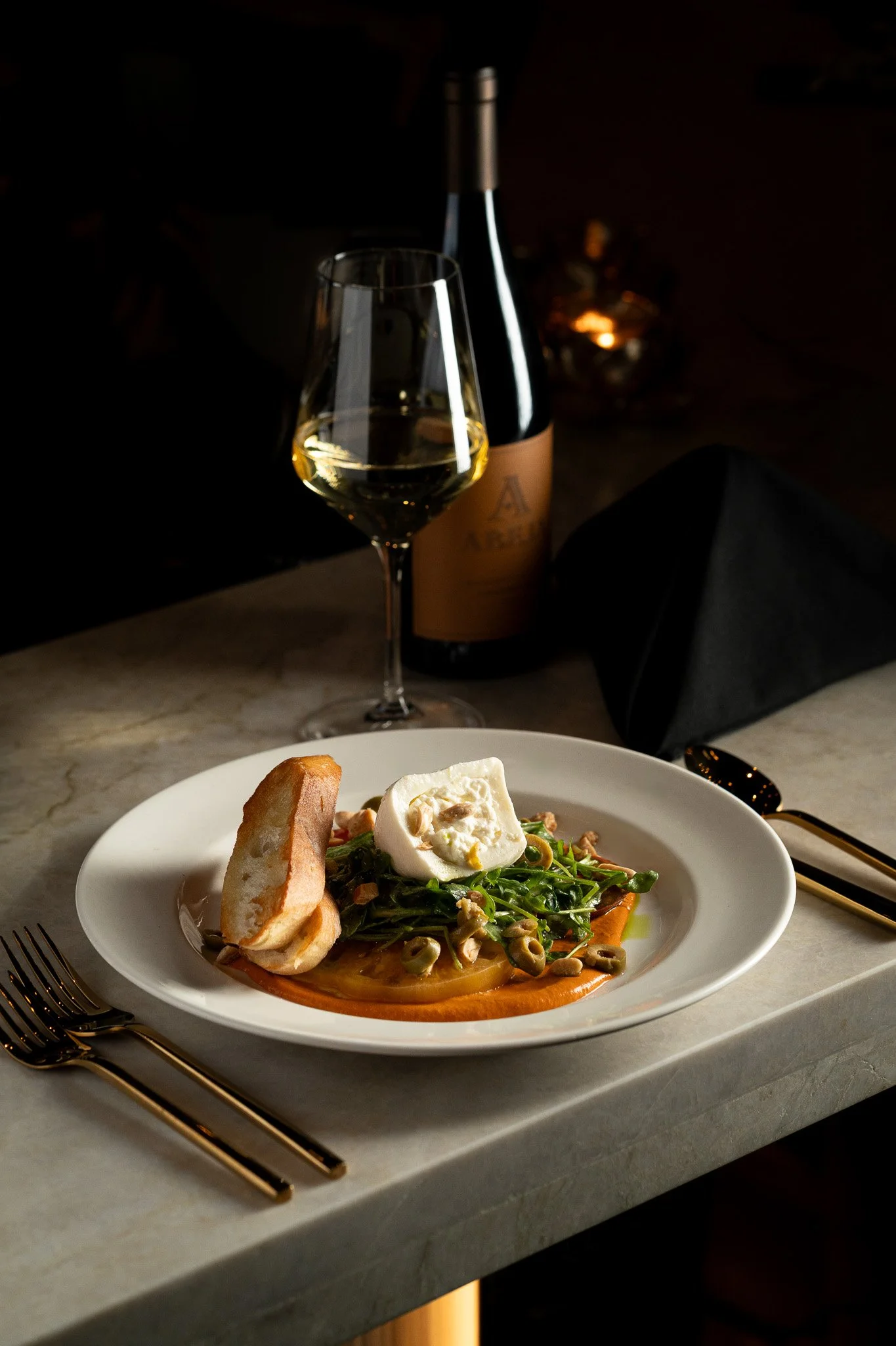 A plated salad with greens, goat cheese, and sliced vegetables, a piece of bread, a glass of white wine, and a bottle of wine on a marble table, with gold utensils and dark background.