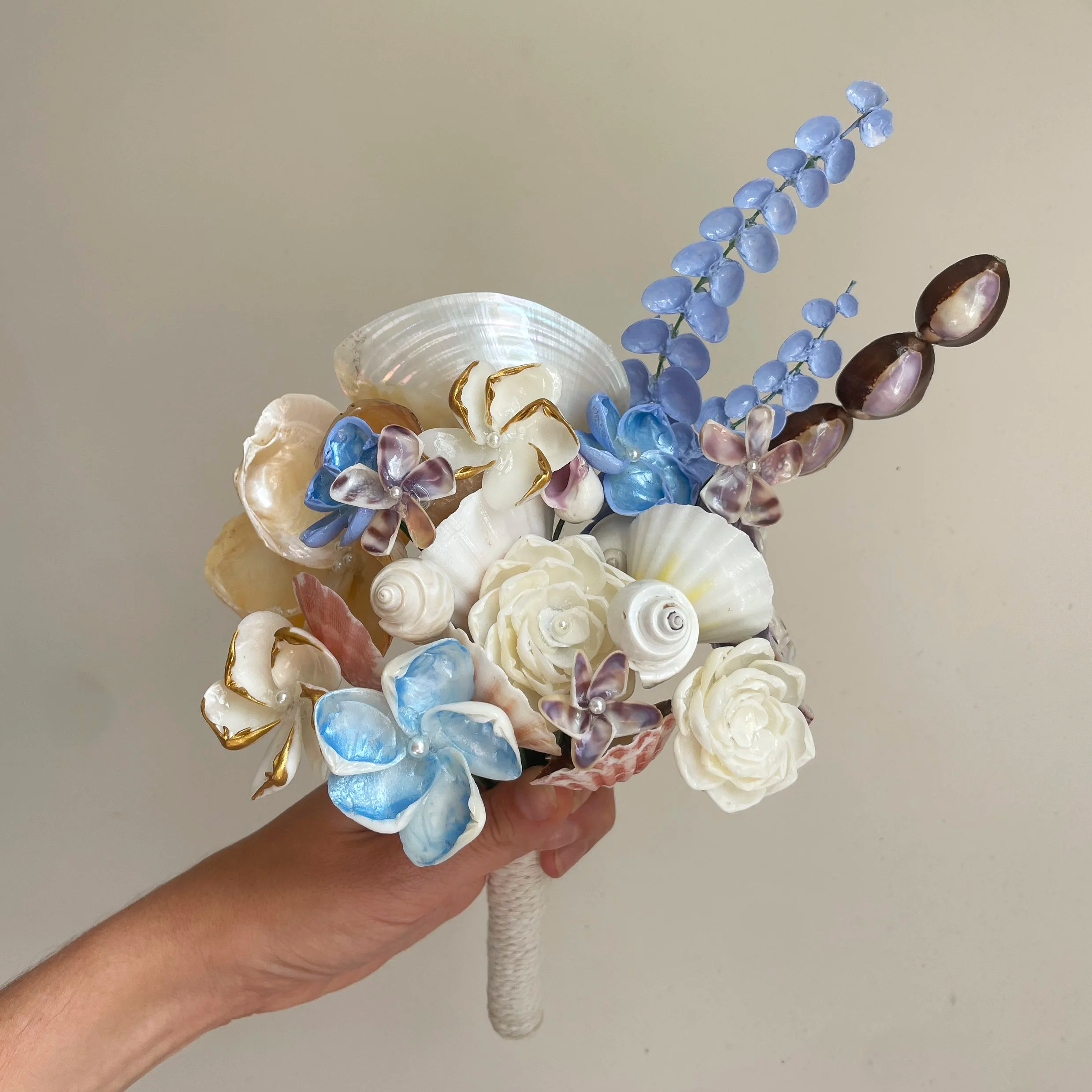 Close-up of a hand holding a decorative bouquet made of seashells and artificial flowers, with a plain beige background.