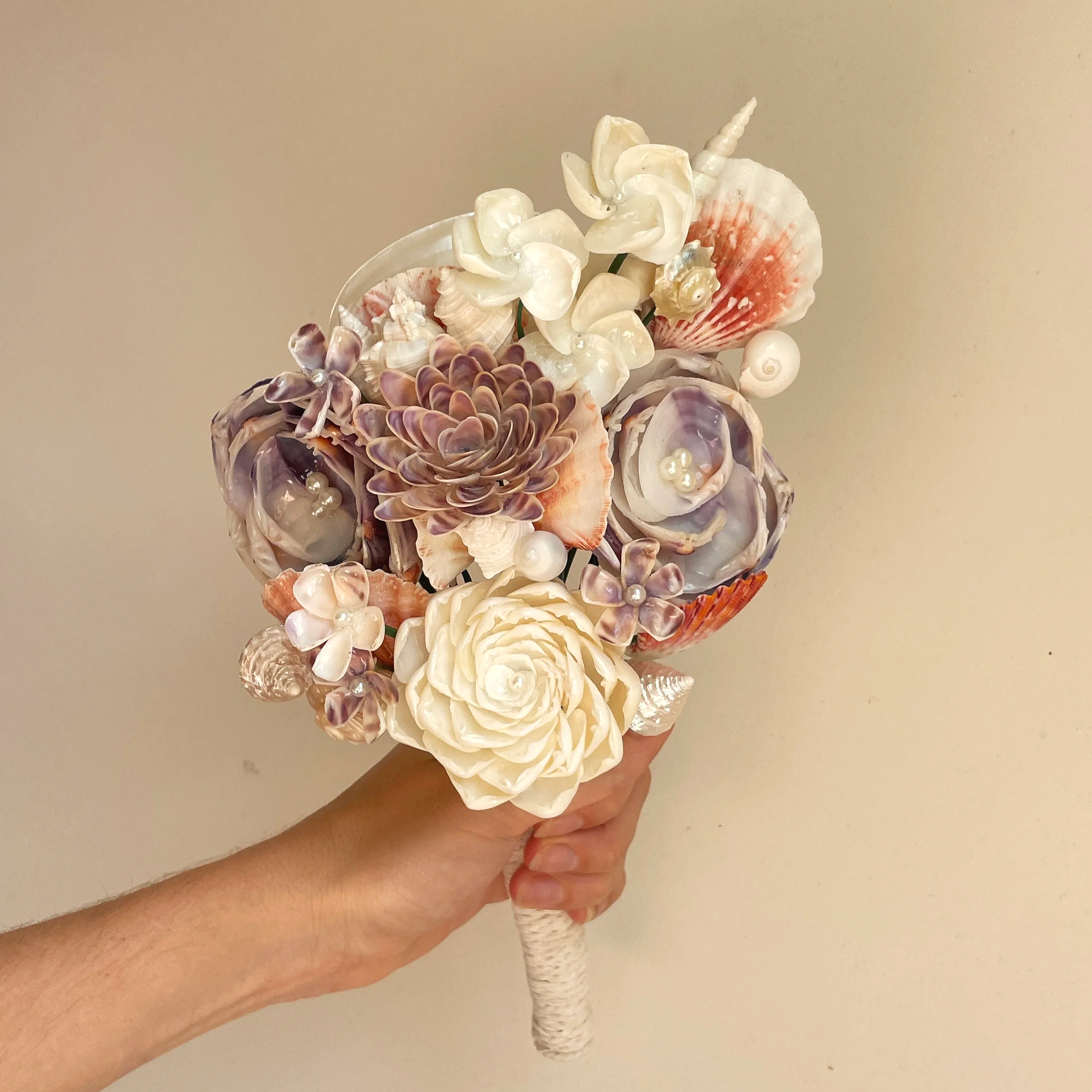 a bouquet made of soft pastel seashells being held by a hand against a white backdrop
