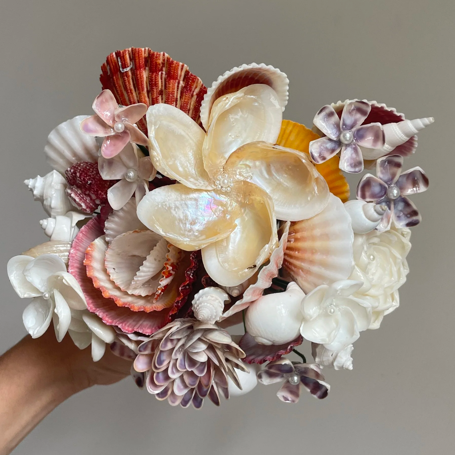 Bouquet of seashells and artificial flowers with pearl accents, held by a hand against a plain background.