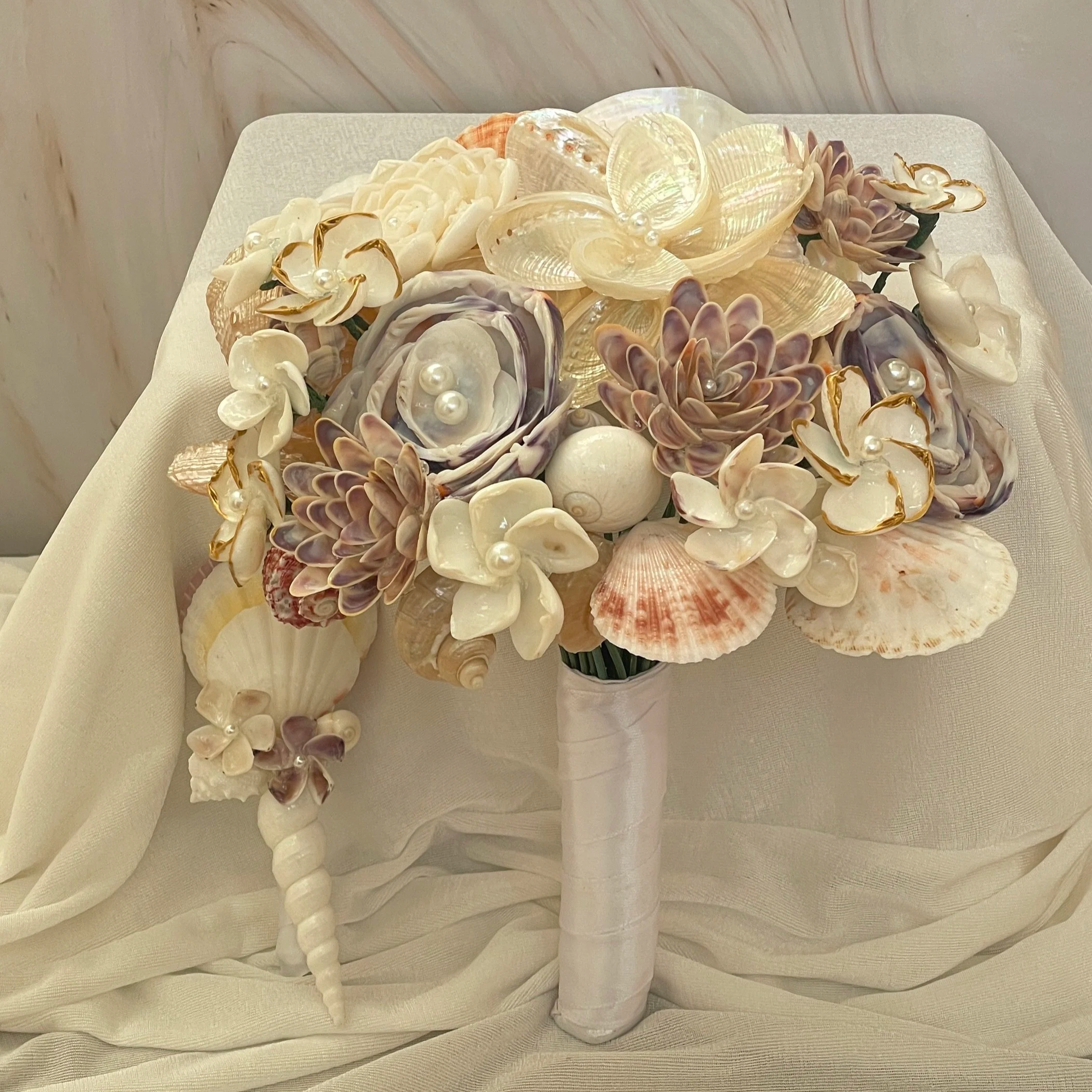 A decorative bouquet of seashells, flowers, and pearls arranged on a white cloth.