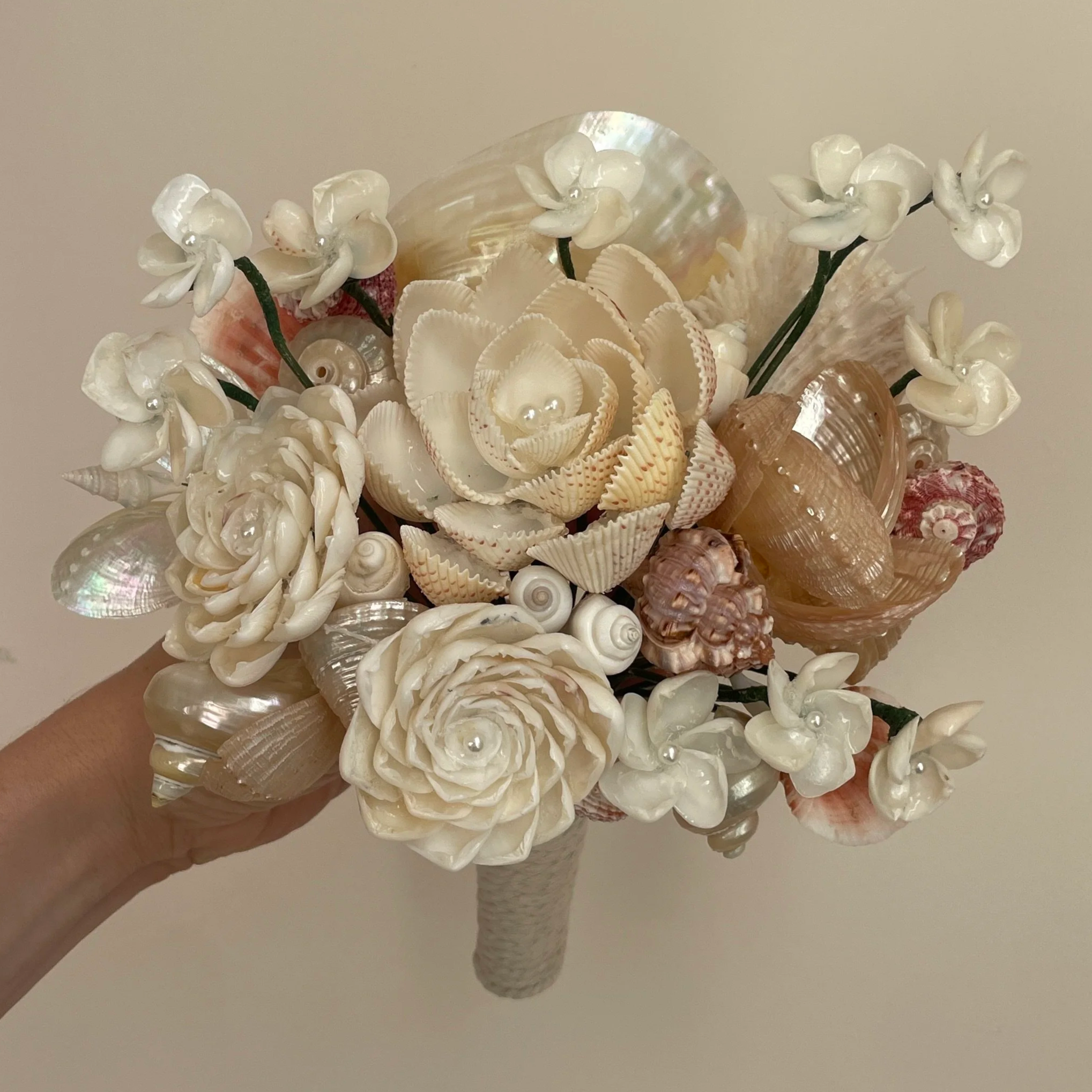 A floral arrangement involving seashells, white flowers, and pearl accents held by a hand, with a plain beige background.