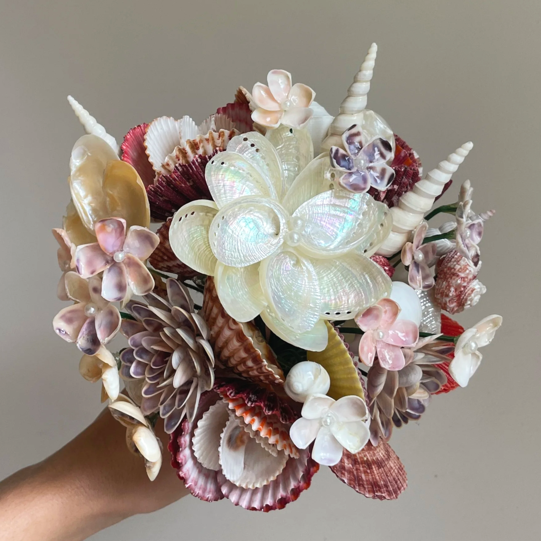 A bouquet made of various seashells, including spiral, scallop, and clam shells, arranged with pink and purple flowers, held by a hand against a plain gray background.