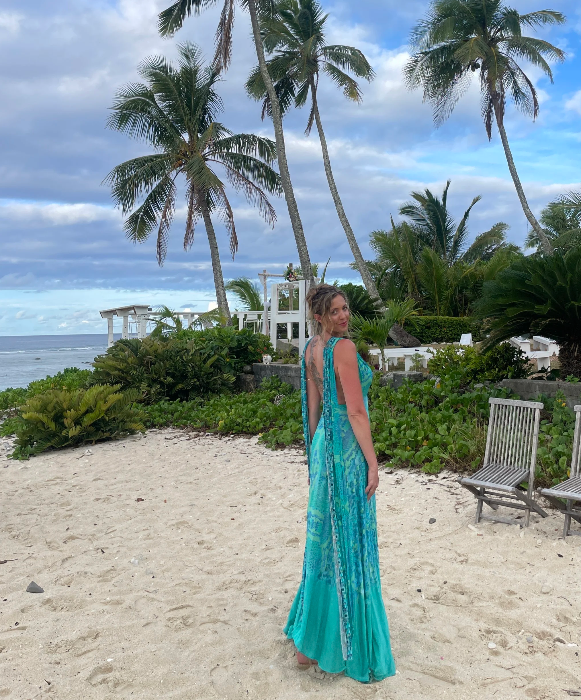 A woman in a long turquoise dress standing on a sandy beach with palm trees and chairs in the background.