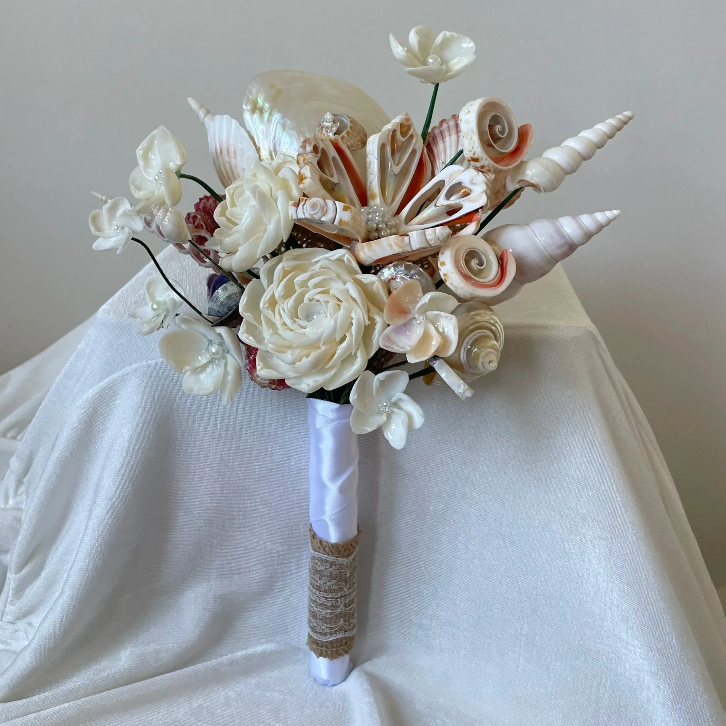 A bouquet made of seashells assembled to resemble flowers, wrapped in white satin ribbon, displayed on a draped white fabric background.