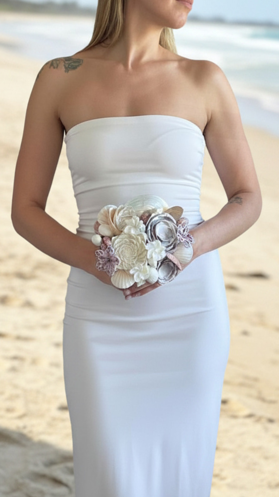 woman in white strapless dress holding a seashell and flower bouquet at the beach.