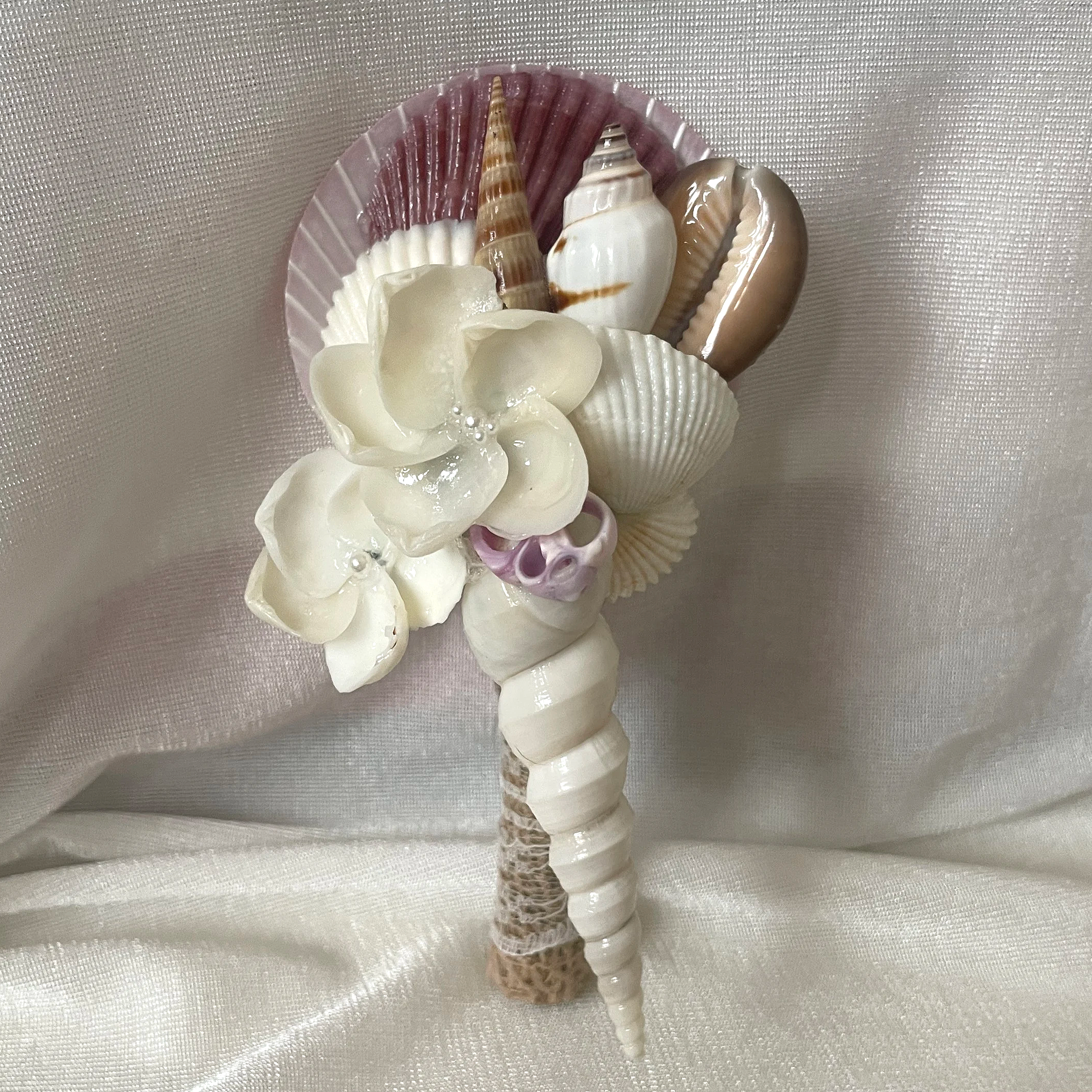 A bouquet made of various seashells and white artificial flowers against a satin fabric background.