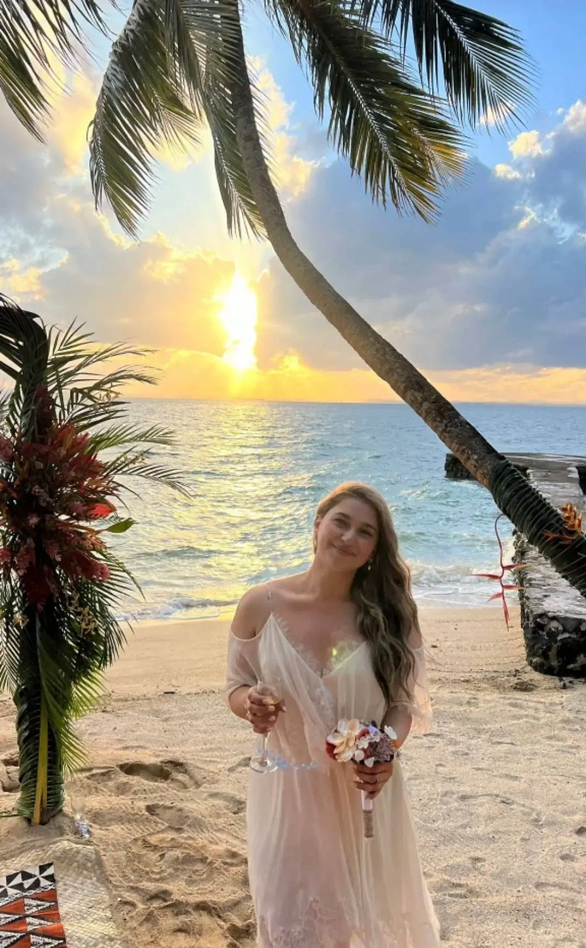 A woman in a light pink dress holding a flower bouquet and a glass of wine, standing on a beach at sunset with palm trees and the ocean in the background.