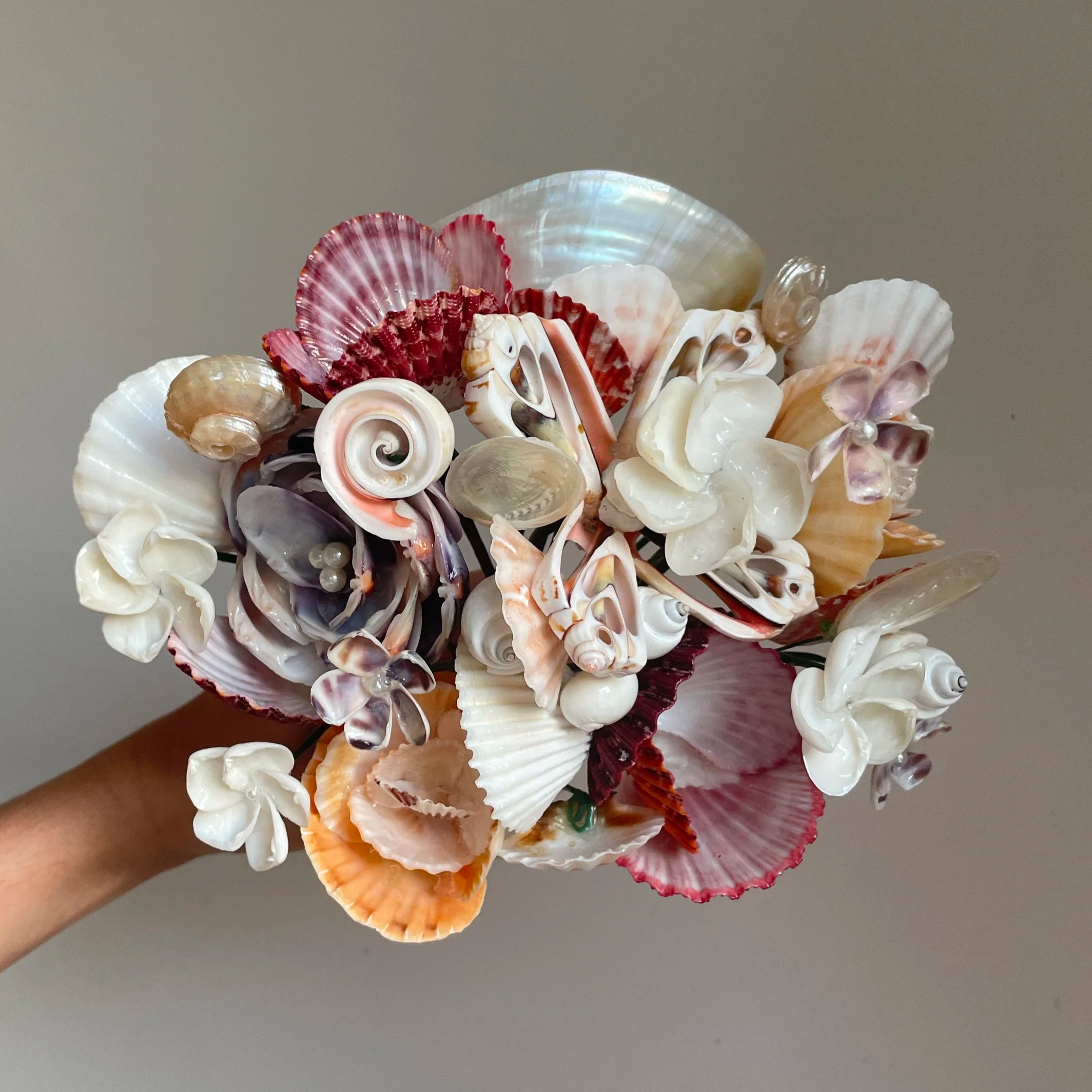 A bouquet of seashells held in a hand, featuring various shapes, sizes, and colors, including pink, white, and beige shells, against a plain gray background.