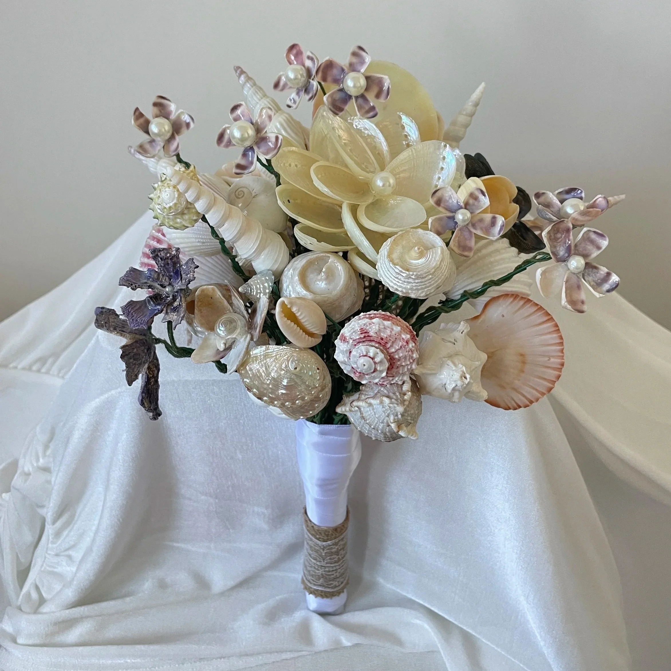 A bouquet of seashells arranged in a vase with pearl accents, placed on a white satin surface.