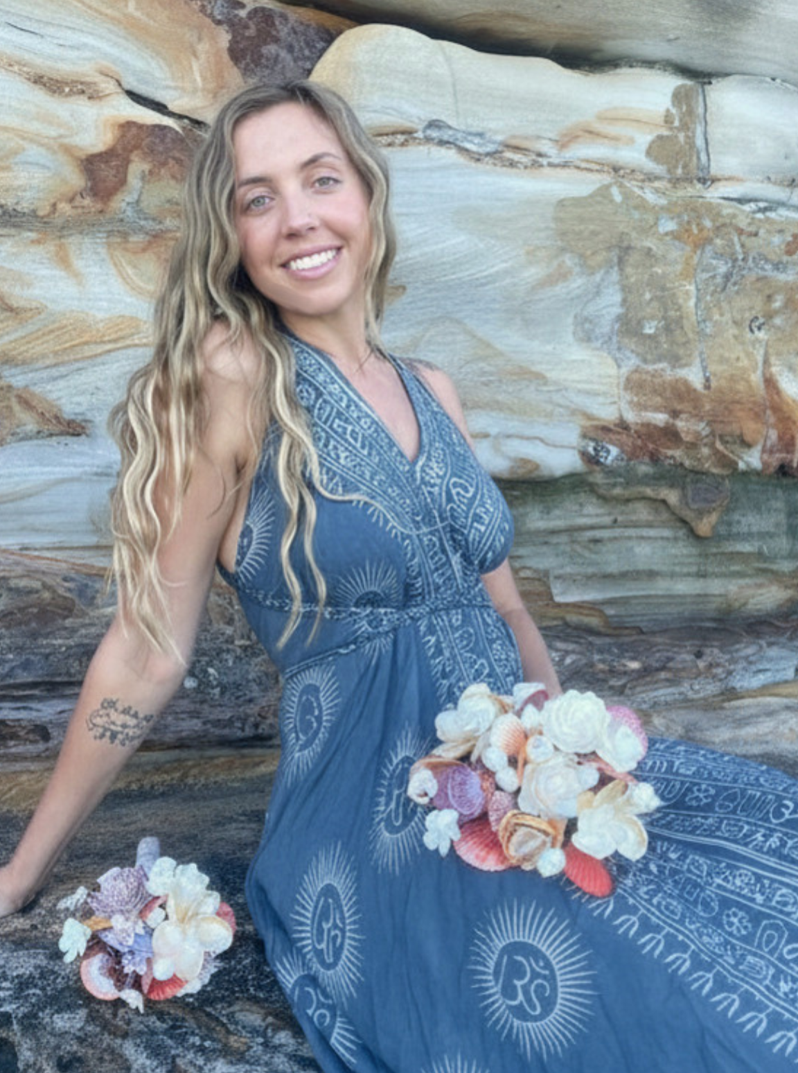 A woman with long wavy blonde hair smiling and sitting against a rock wall, holding shells and flowers, wearing a blue patterned dress.