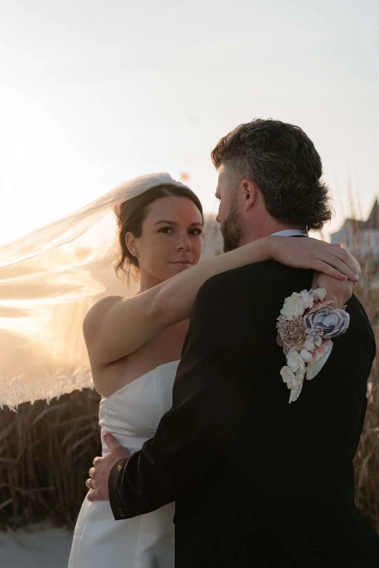 A bride and groom dancing outdoors at sunset, with the bride wearing a white dress and veil, and the groom in a black suit with a floral boutonniere.