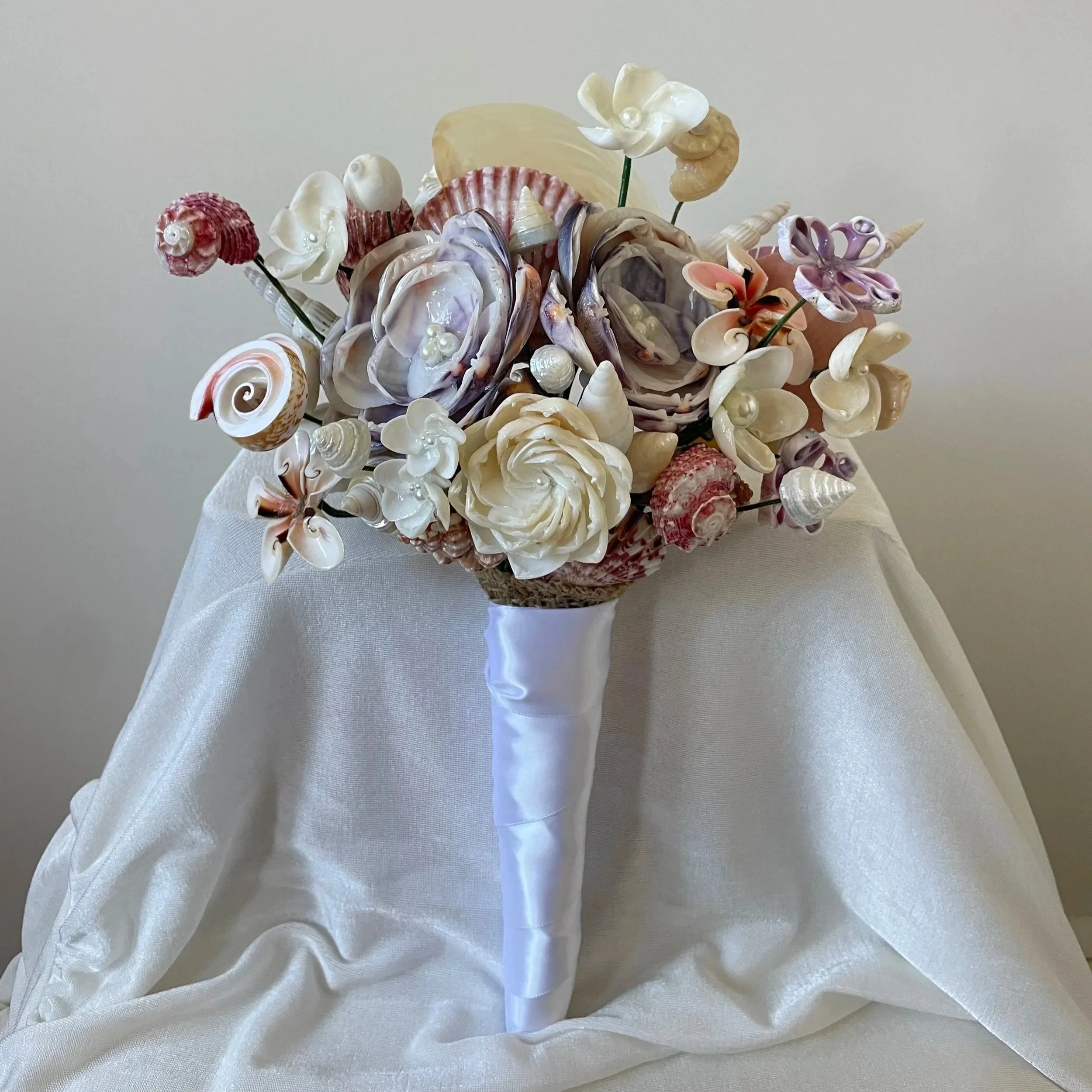 Flower bouquet made of seashells and pearl accents, wrapped in satin ribbon, resting on ivory fabric.
