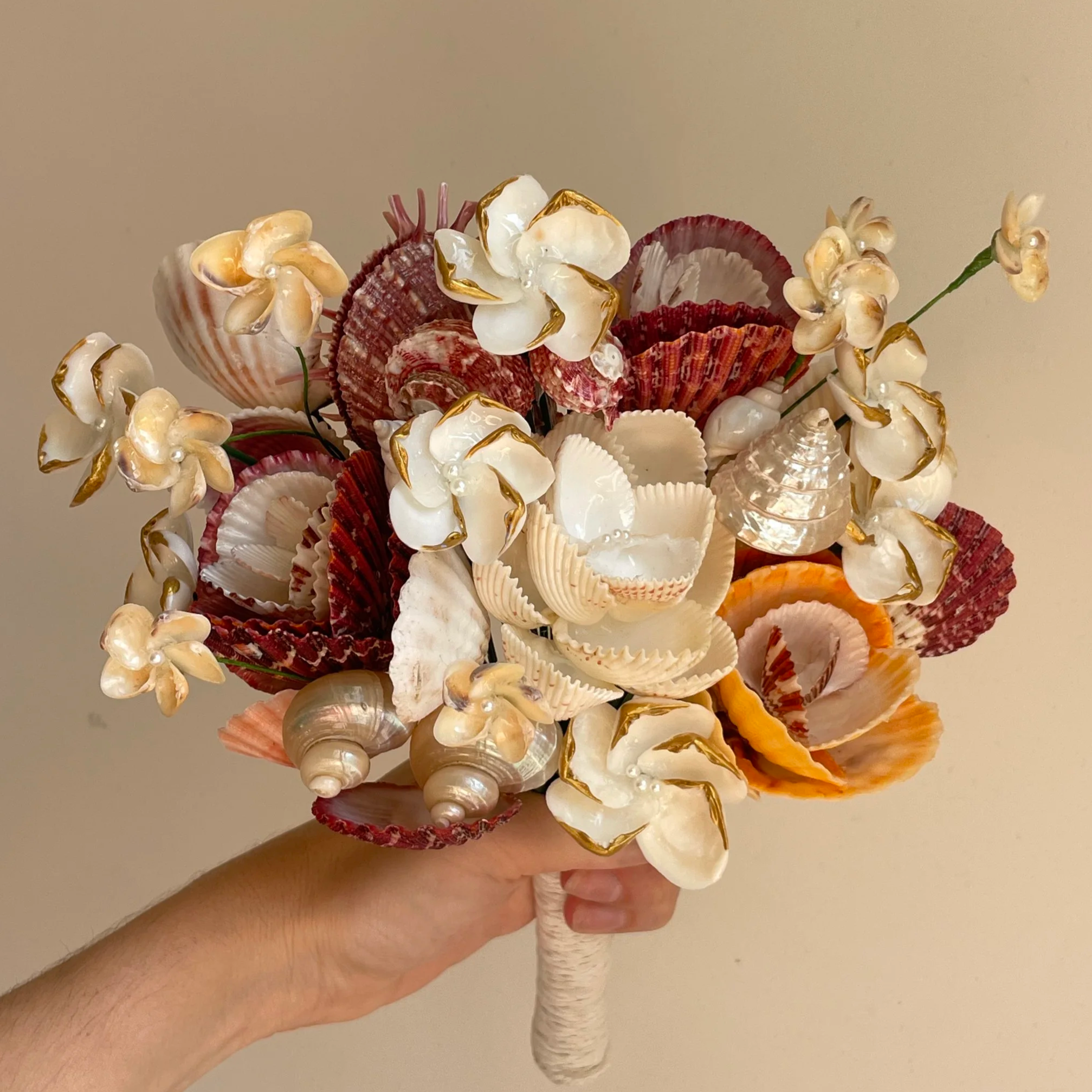 A bouquet of red and gold seashells being held by a hand in front of a plain white backdrop