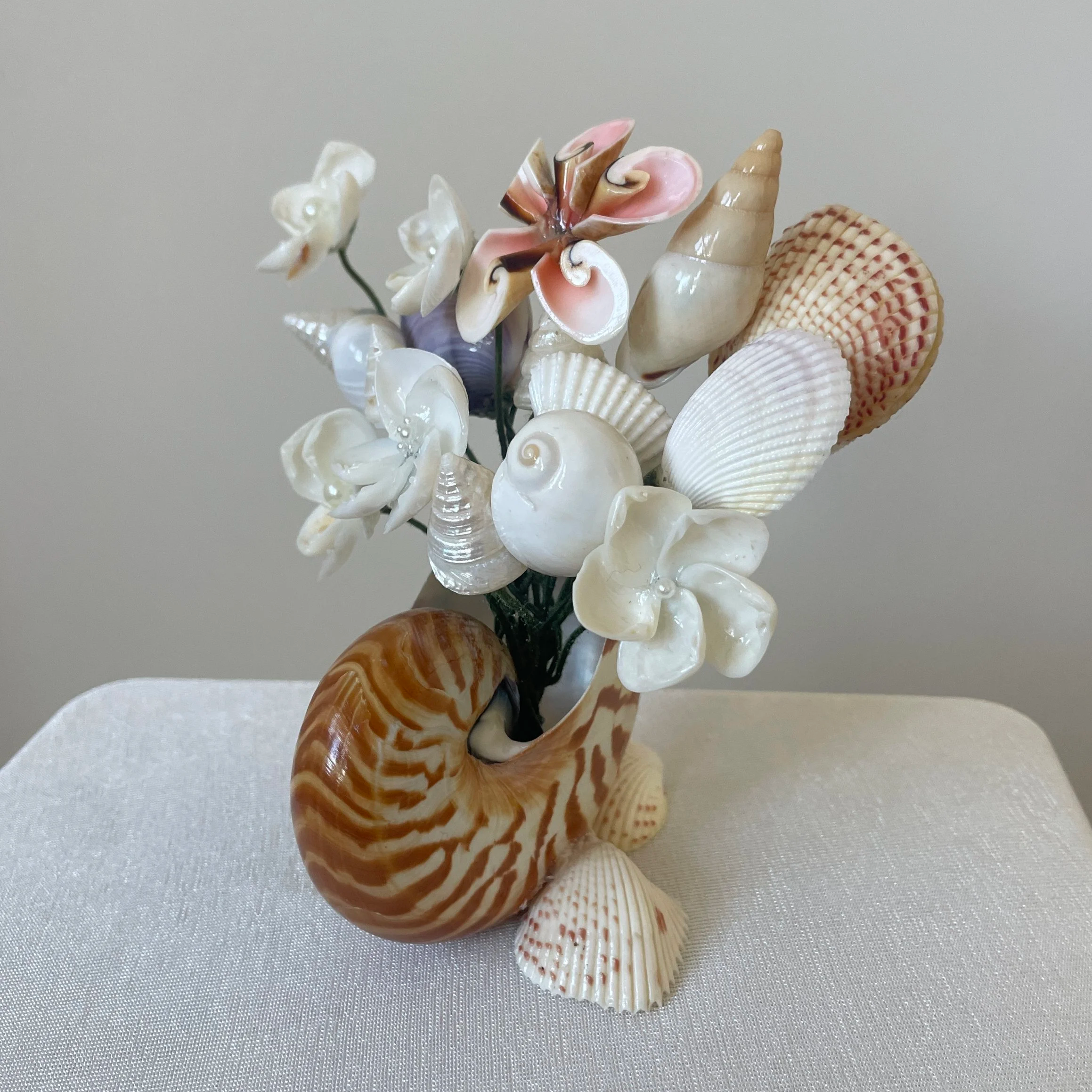 Decorative arrangement of various seashells and artificial flowers in a striped nautilus shell vase on a white table
