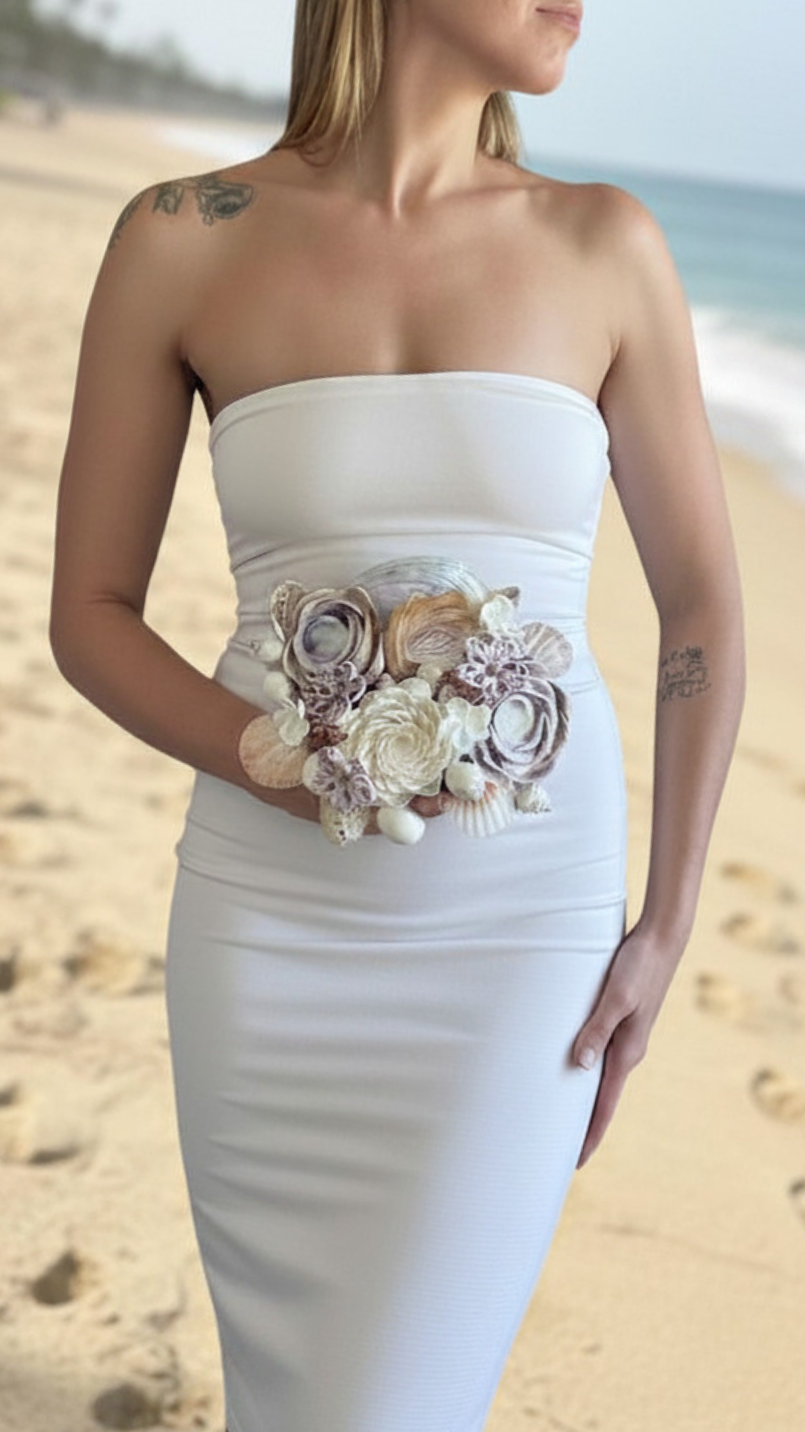 A woman in a strapless white dress with a seashell and flower embellishment on the waist, standing on a beach.