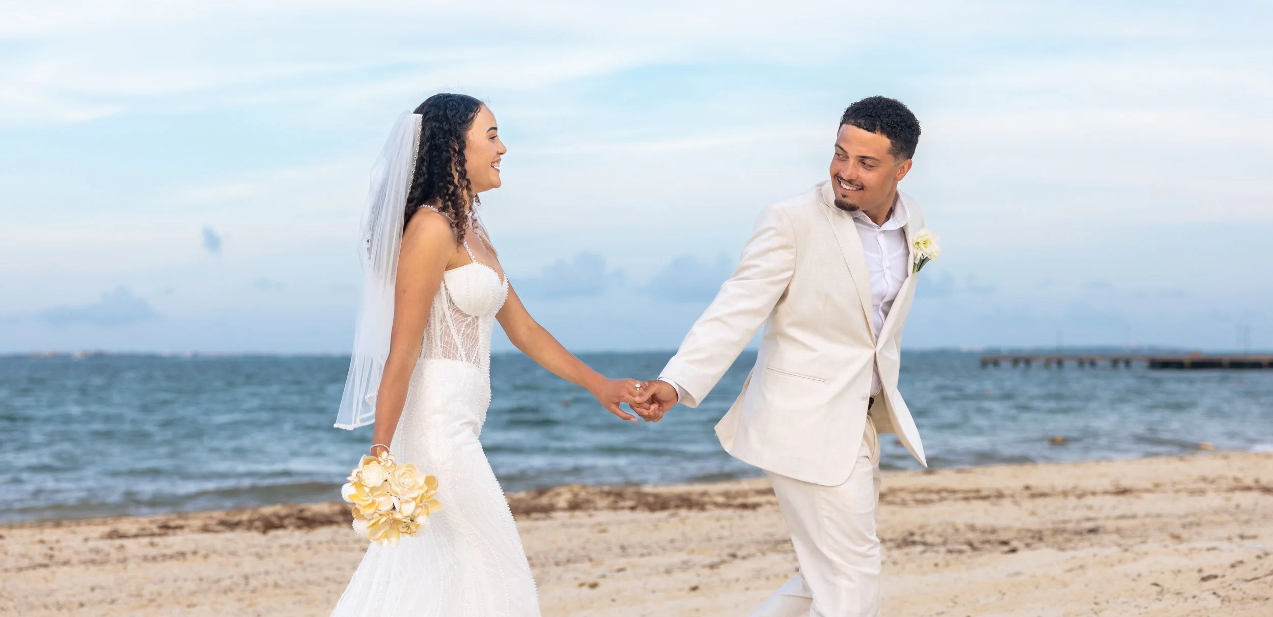 A bride and groom on the beach, holding hands, smiling, with the ocean and cloudy sky in the background, dressed in wedding attire.