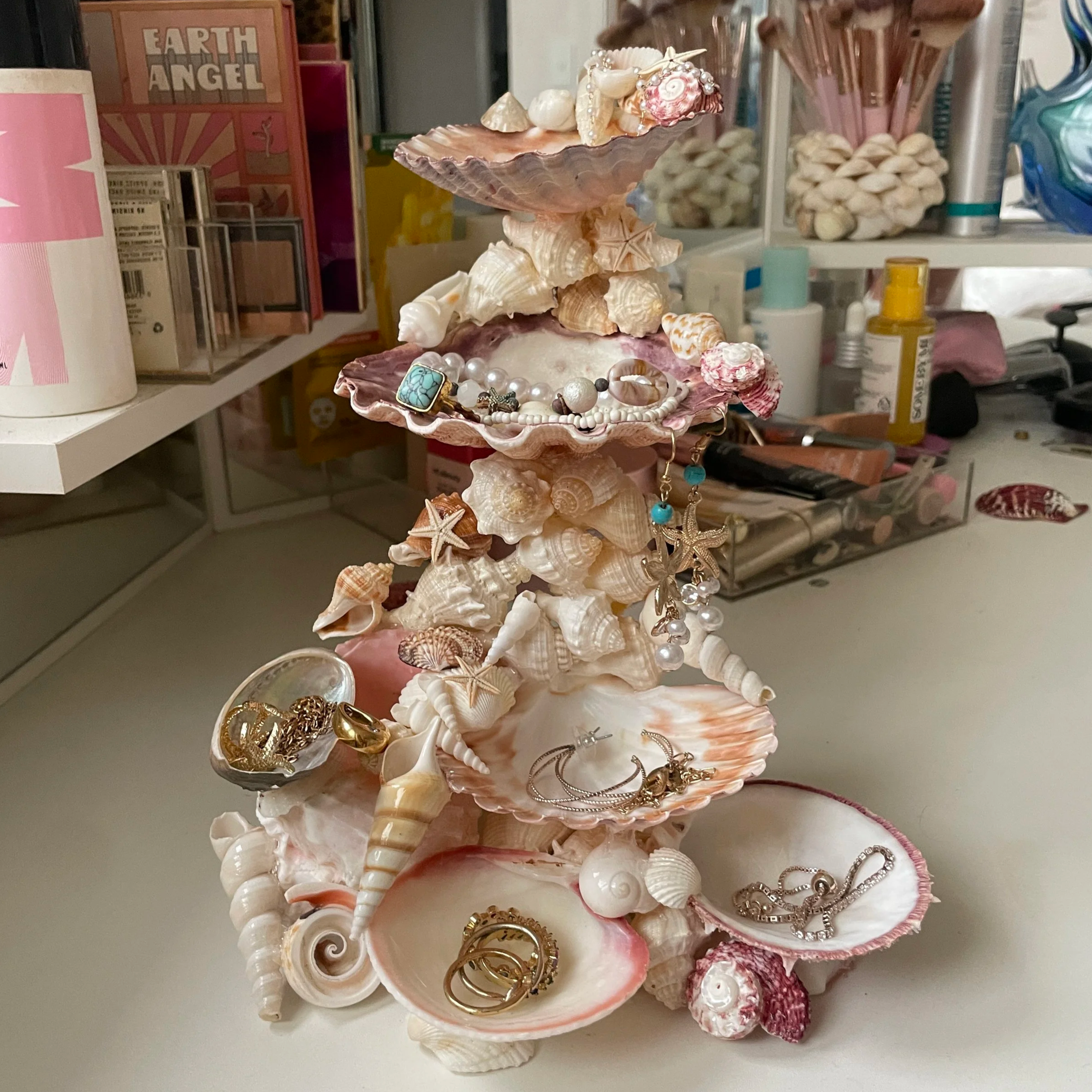 A jewellery tower made entirely of seashells on a vanity, it has draping gold jewellery on it, and make up in the back ground 
