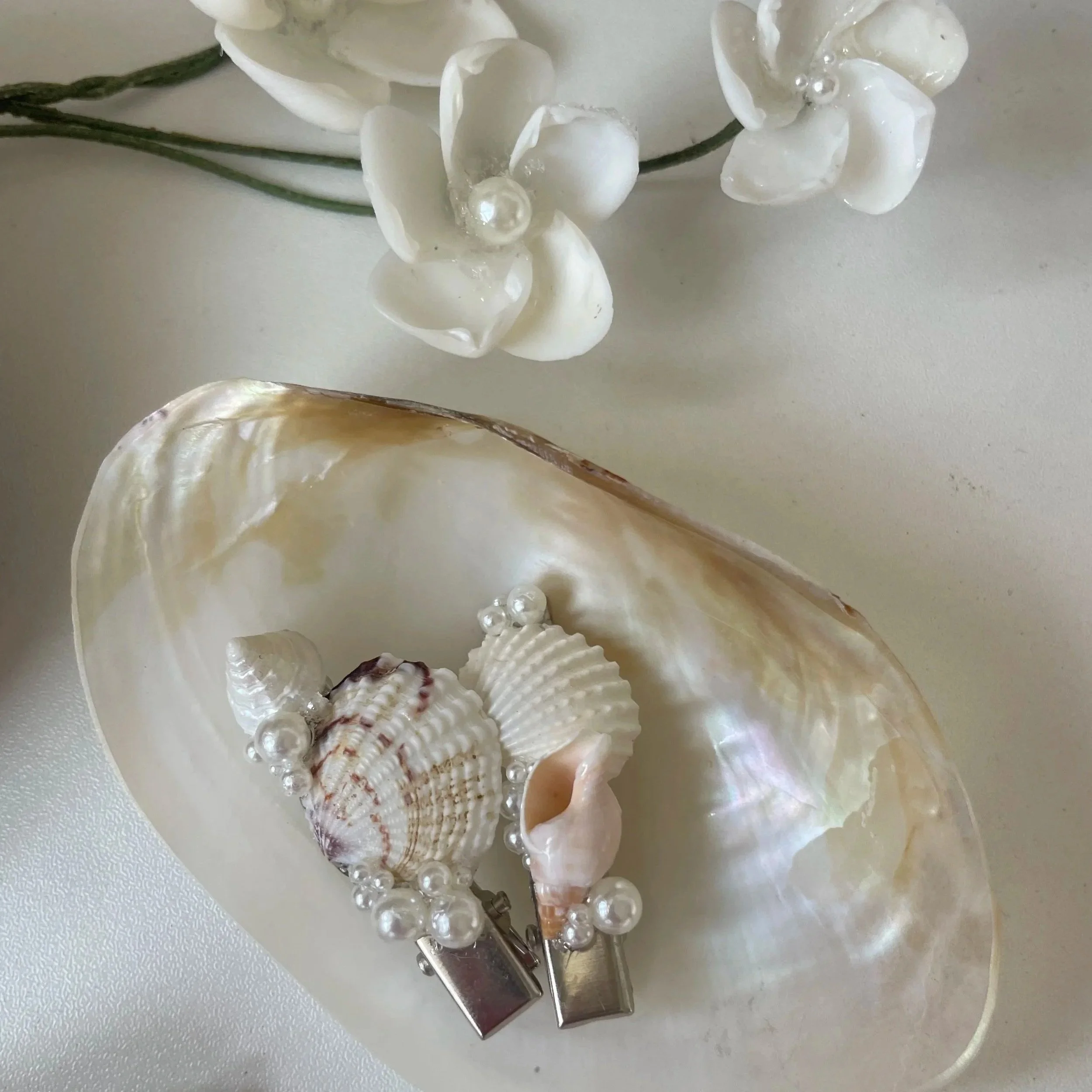 A large seashell with smaller seashells and pearl decorations inside, and white artificial flowers with pearl accents in the background.