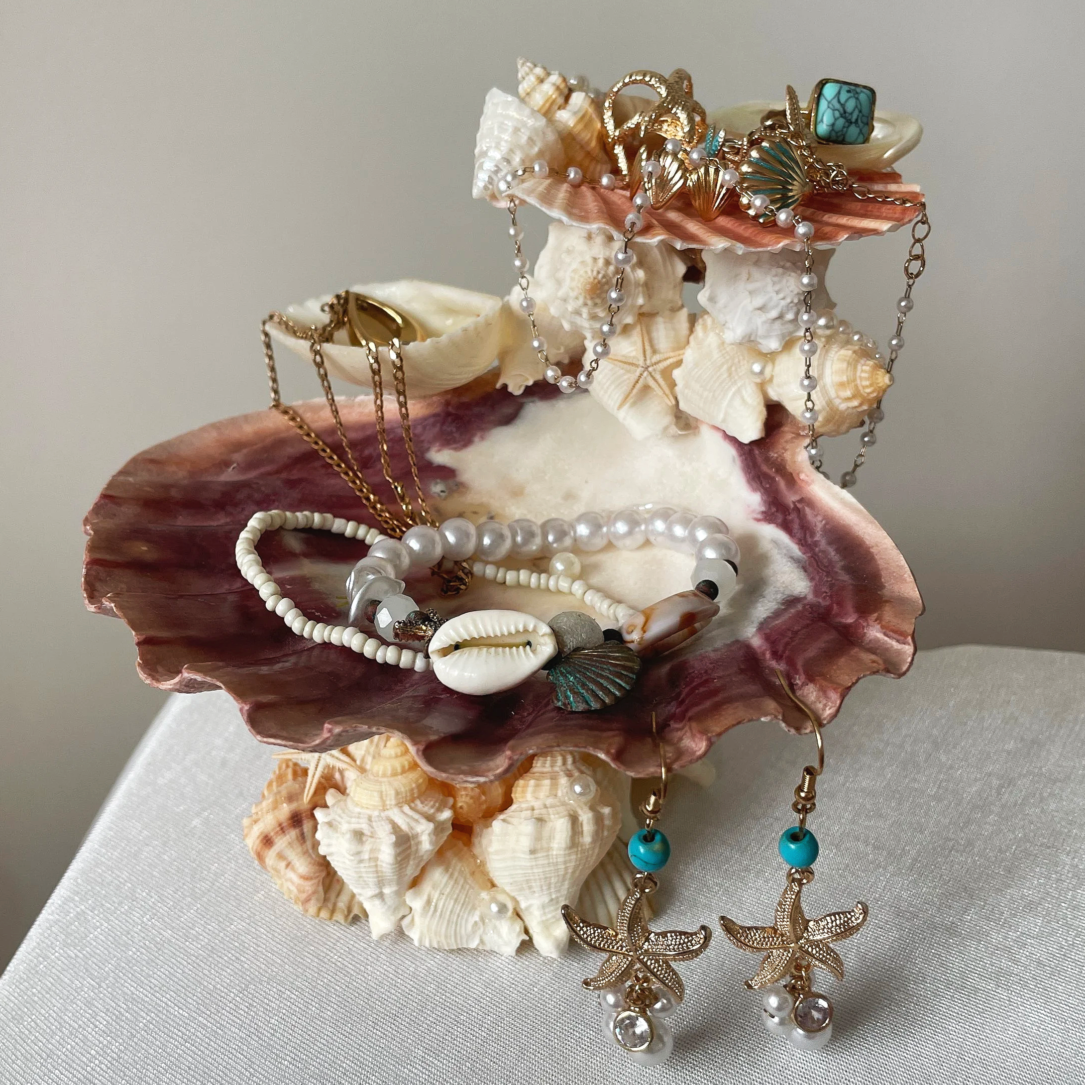A trinket dish made of a lions paw scallop shell and smaller shells, on a white table with a lain background, with gold jewellery on top and draping across it