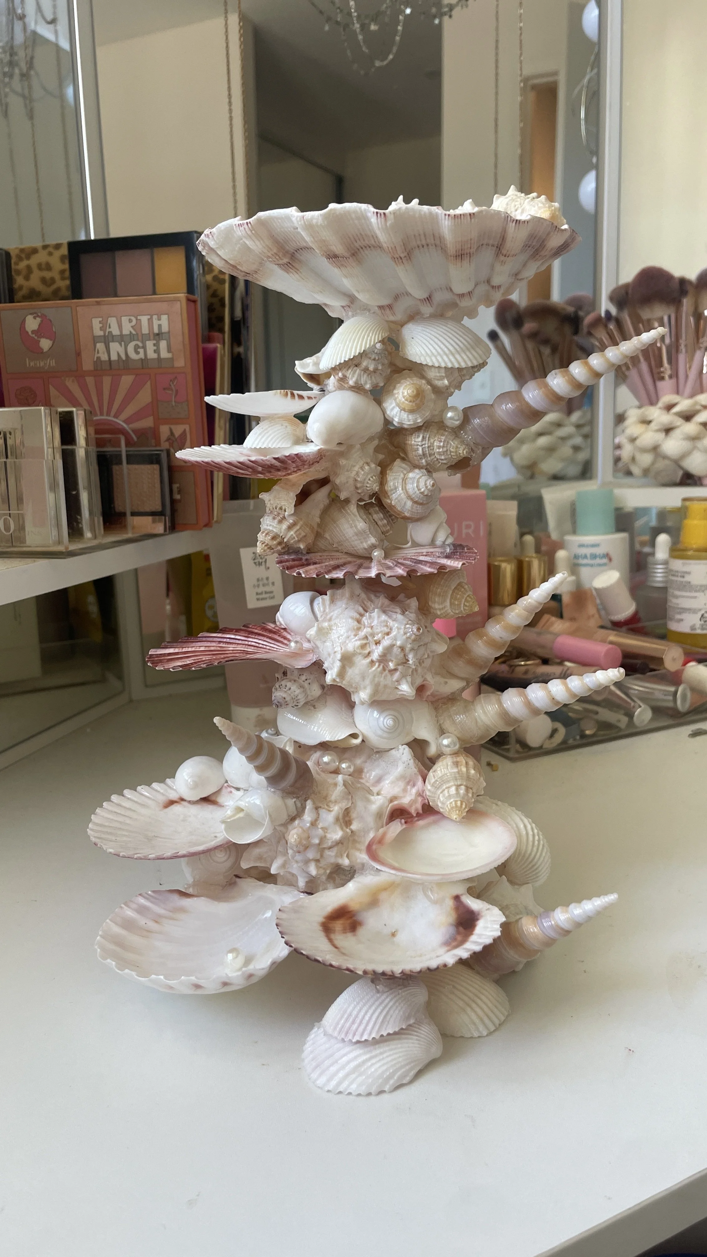 Decorative tower made of various seashells on a white surface, with makeup and skincare products in the background.