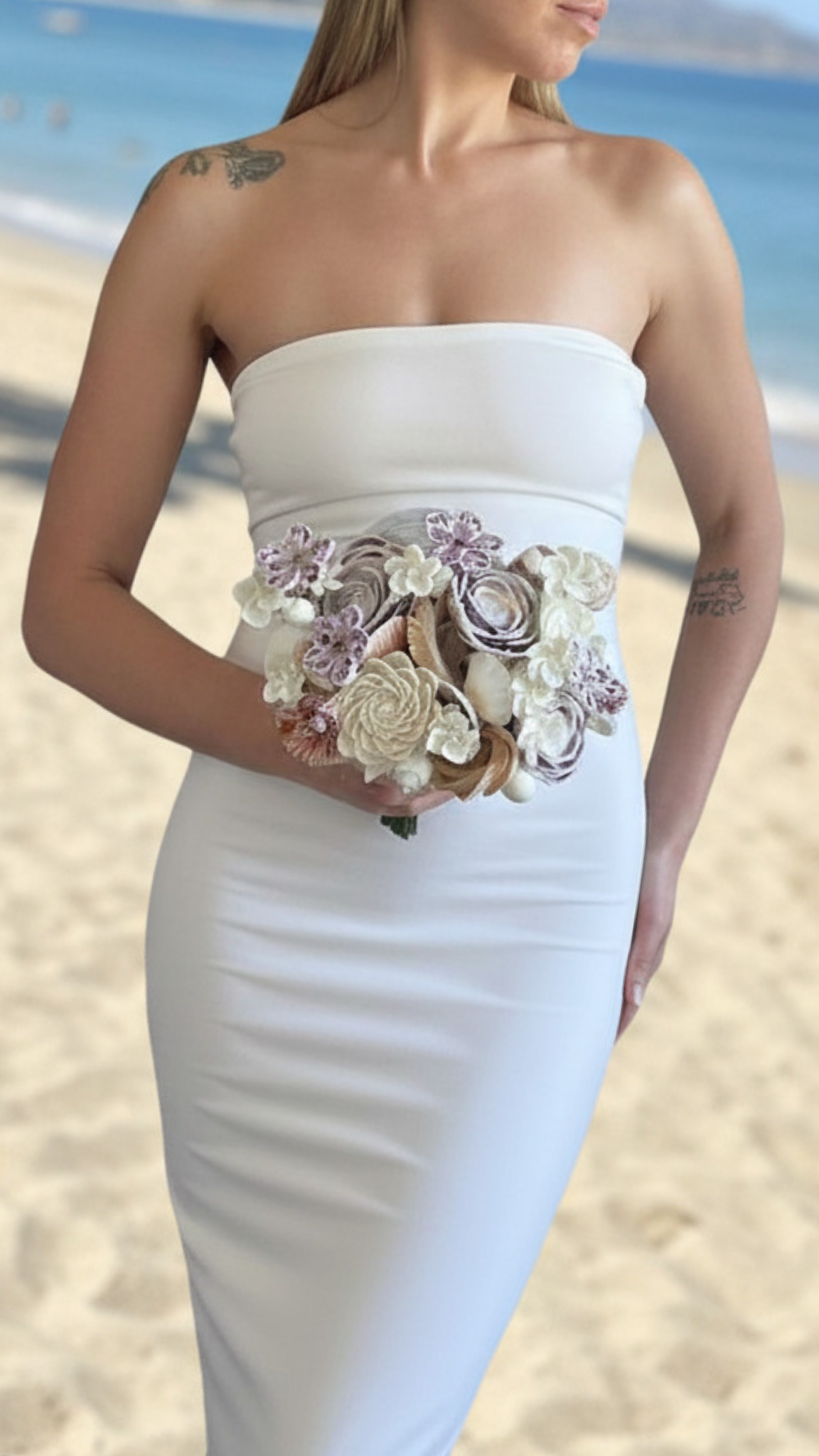 A woman in a strapless white dress holding a bouquet of pastel-colored flowers on a beach.