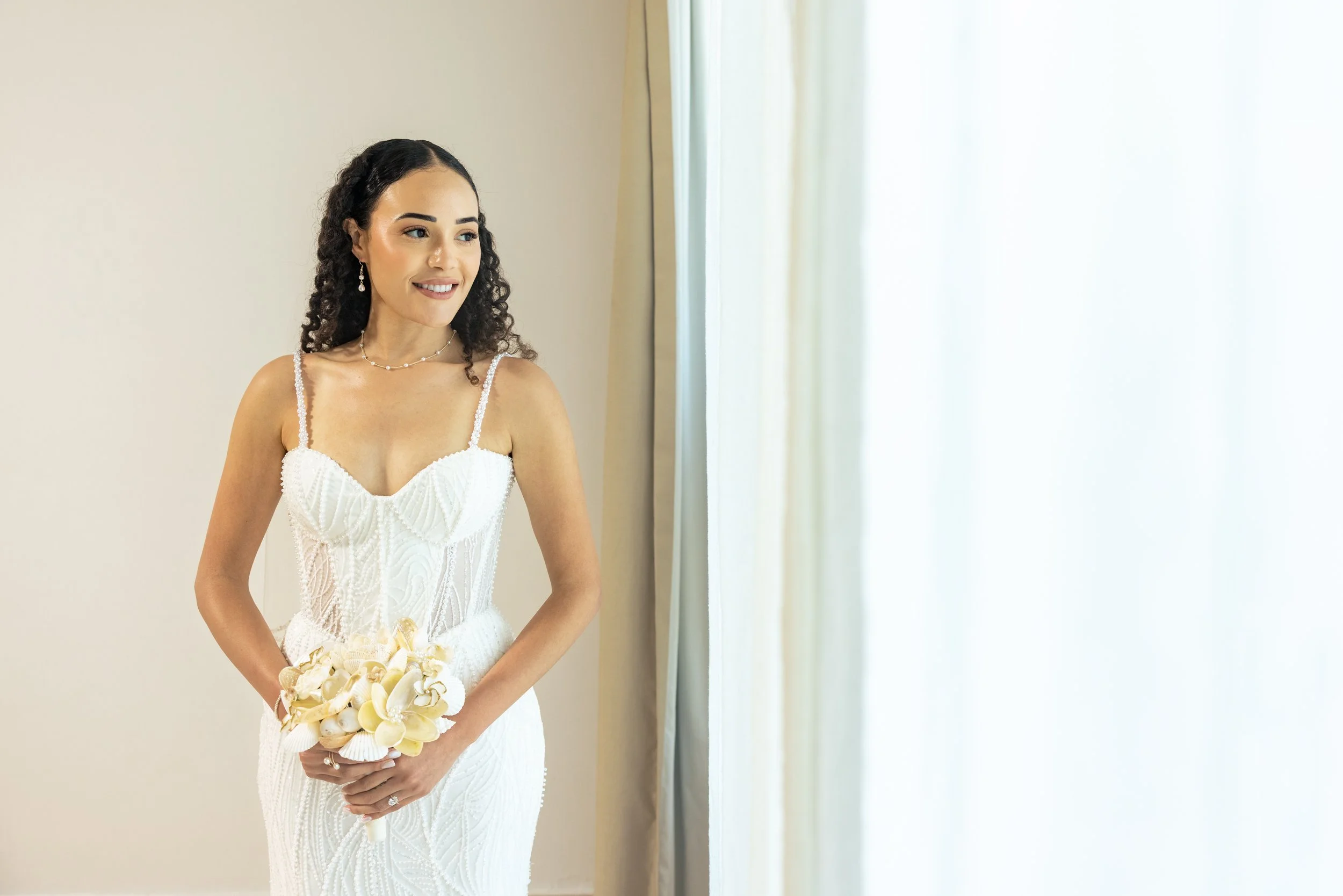 A woman in a white dress holding a bouquet of white and cream flowers, standing near a window with light-colored curtains.