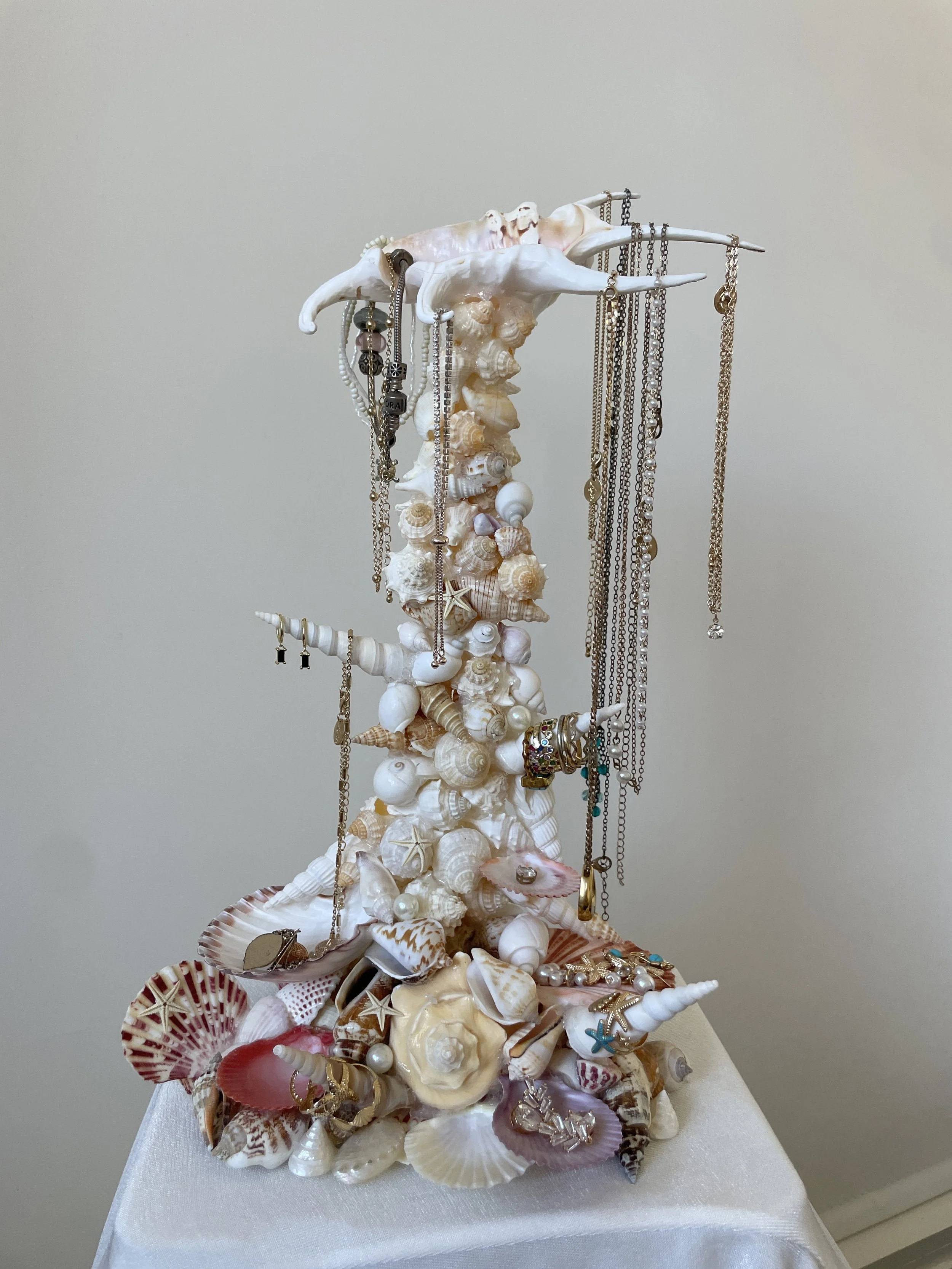 Display of jewelry on a decorative stand made of seashells, beads, and starfish. The jewelry includes necklaces, rings, and earrings, arranged on and around the seashell stand.