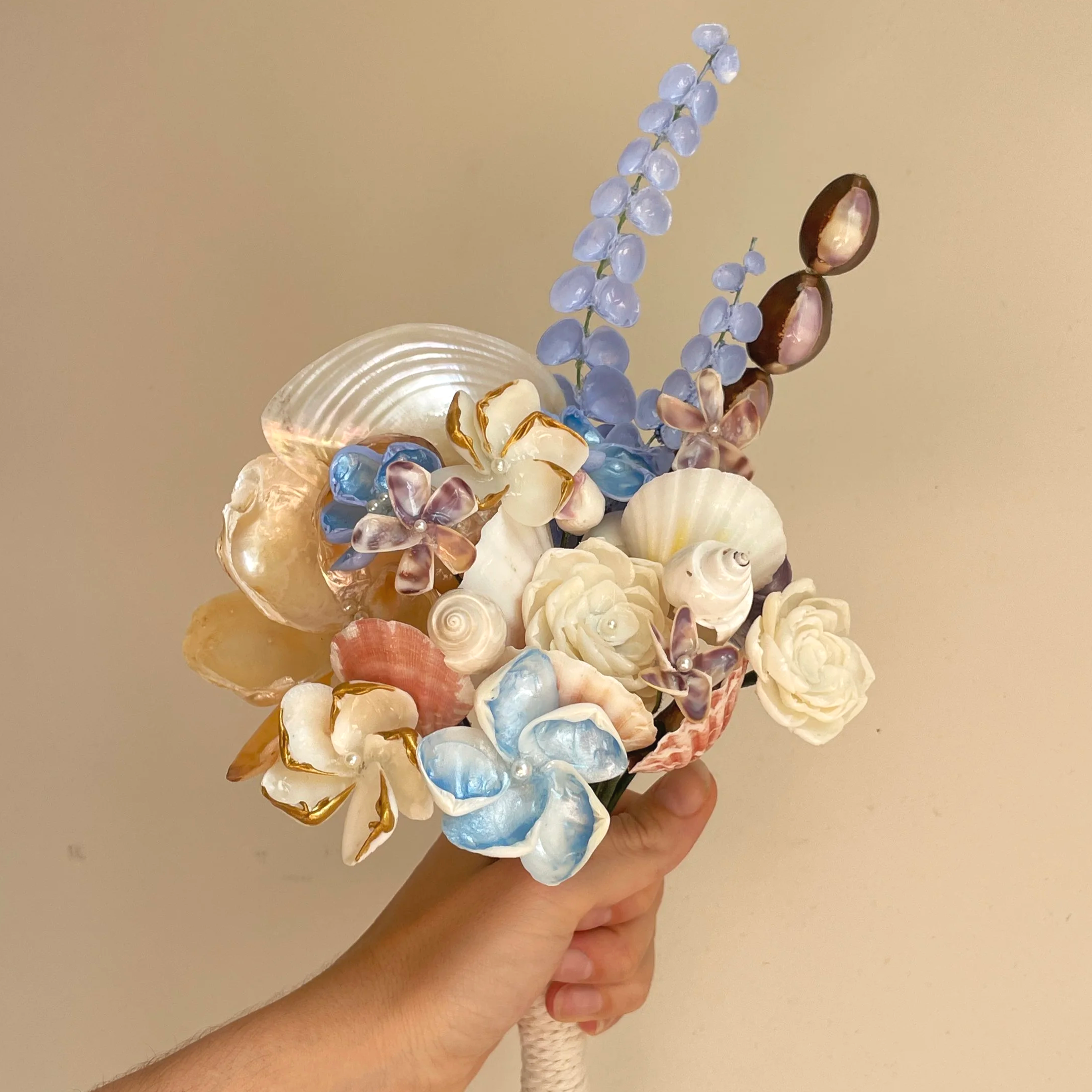 A bouquet made of blue, gold and silver seashells being held by a hand on front of a plain white backdrop