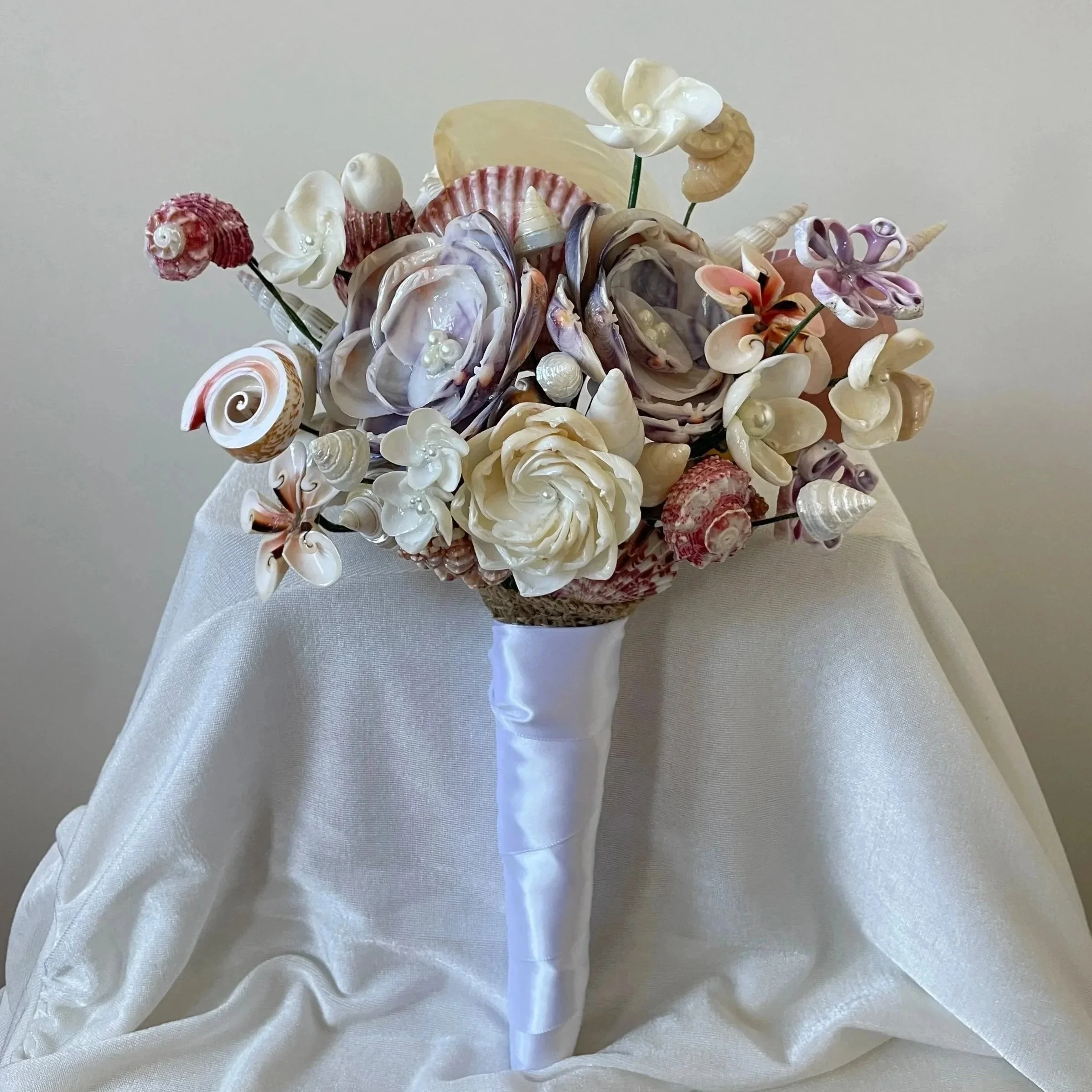A bouquet of seashells arranged to resemble flowers, wrapped with white satin ribbon, placed on a draped white fabric background.
