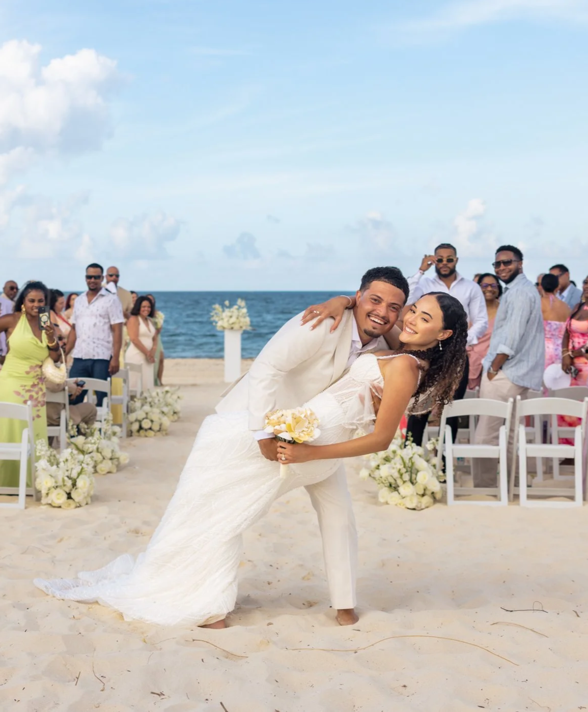 A newlywed couple dressed in wedding attire dancing on a beach, surrounded by friends and family, with the ocean and partly cloudy sky in the background.