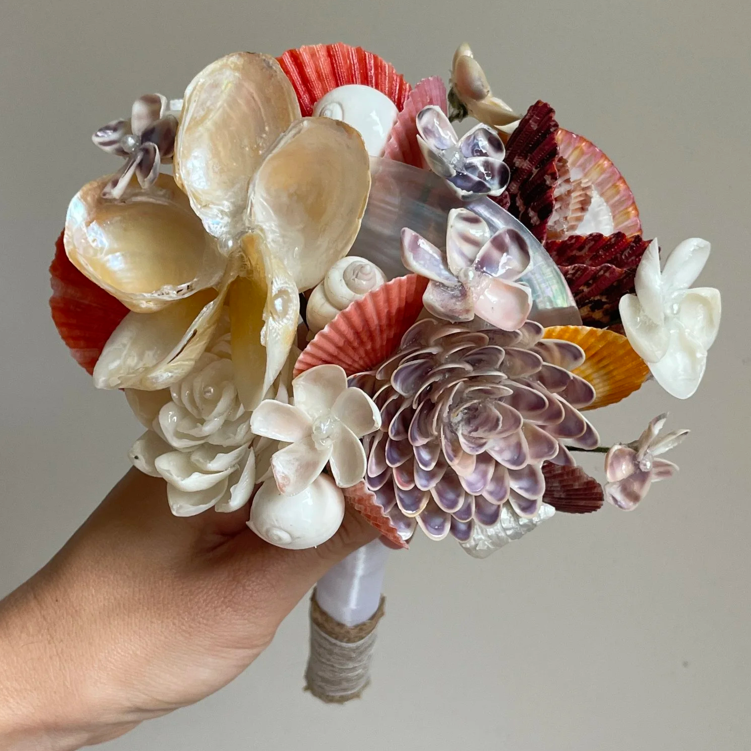 A bouquet of seashells and artificial flowers held by a person's hand, featuring various colorful shells and floral elements.