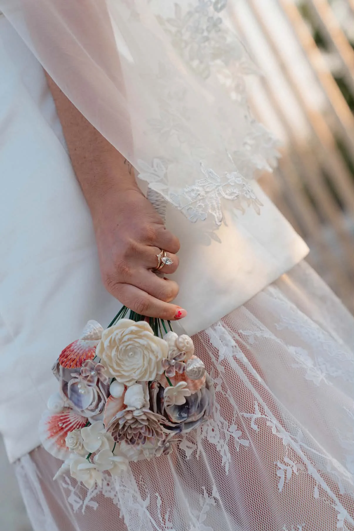 Close-up of a hand holding a bouquet of seashells and flowers, with a bridal lace dress in the background.