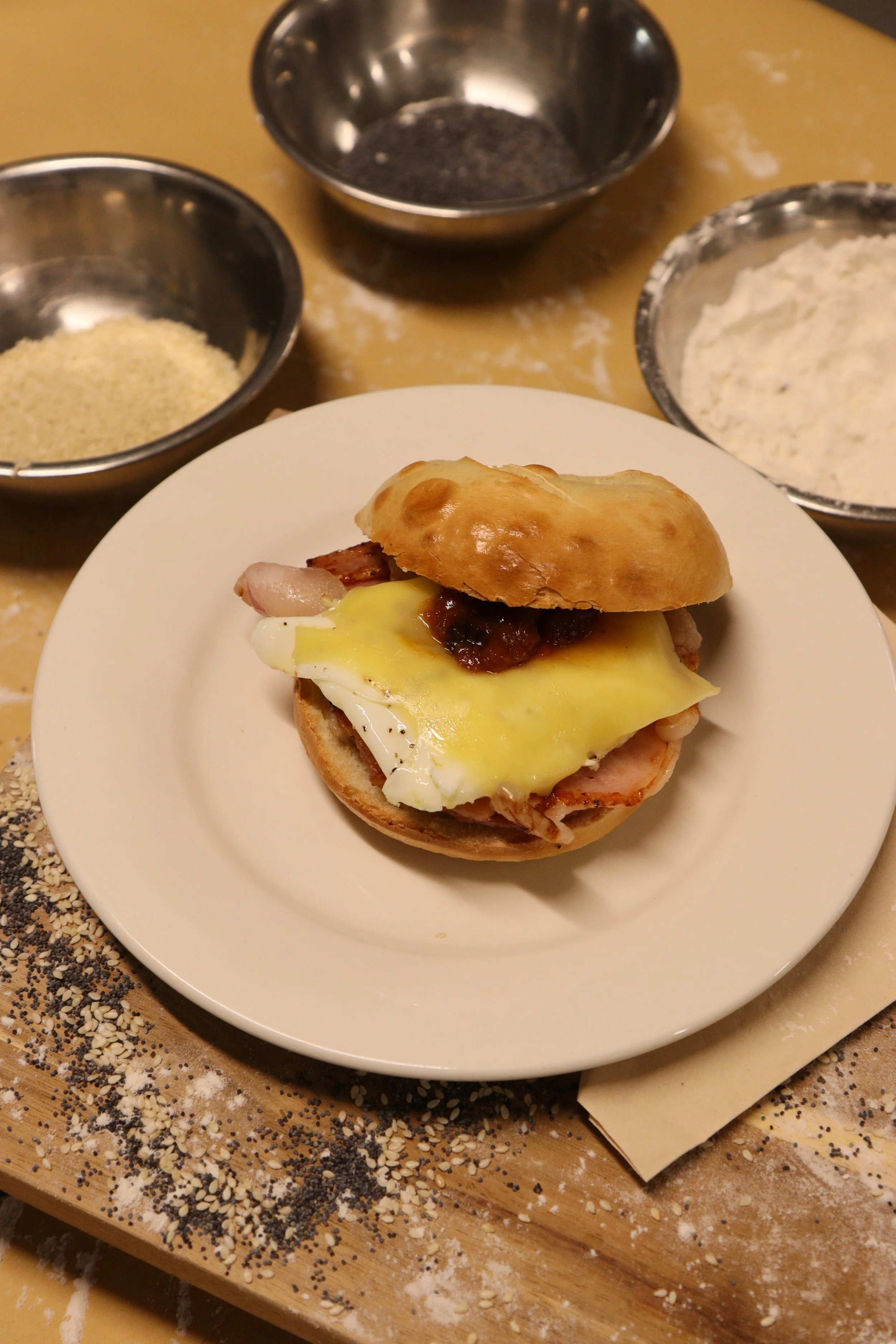 A small sandwich on a white plate with a slice of cheese, bacon, and sauce inside a sandwich bun. In the background, there are three metal bowls with ingredients, including breadcrumbs, poppy seeds, and flour, on a wooden surface with scattered seeds and flour.